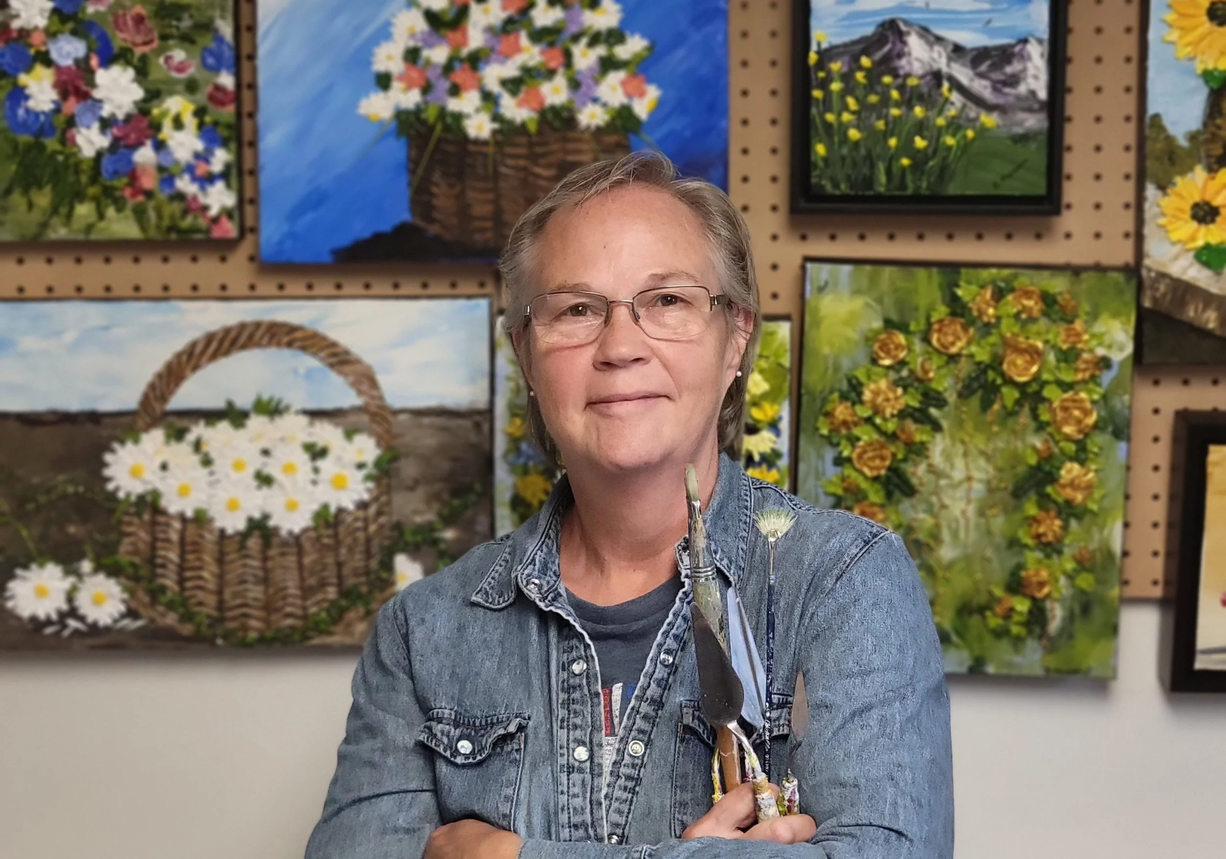 A woman with short gray hair, glasses, and a denim jacket holding paintbrushes, standing in front of a wall with colorful landscape paintings of flowers and mountains.