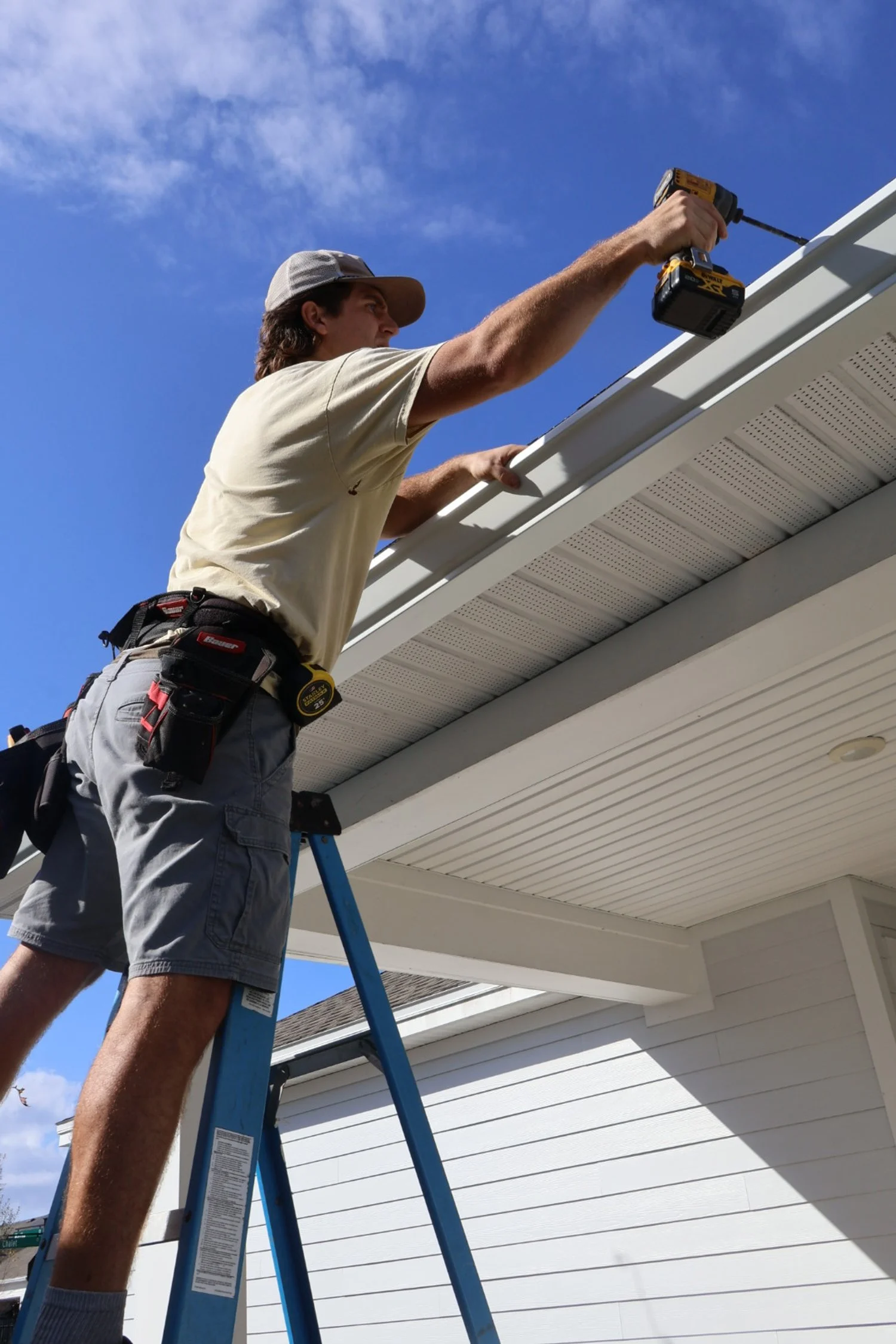 A man standing on a ladder working on the roof of a house, holding a power drill.