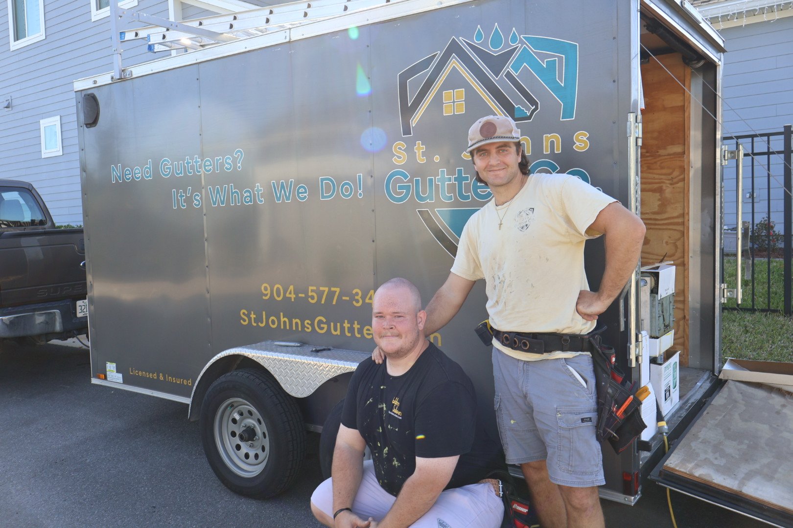 Two men posing in front of a mobile gutter business trailer, one standing and one kneeling, with the trailer displaying the business name St. Johns Gutters, contact information, and slogan 'Need Gutters? It's What We Do!'