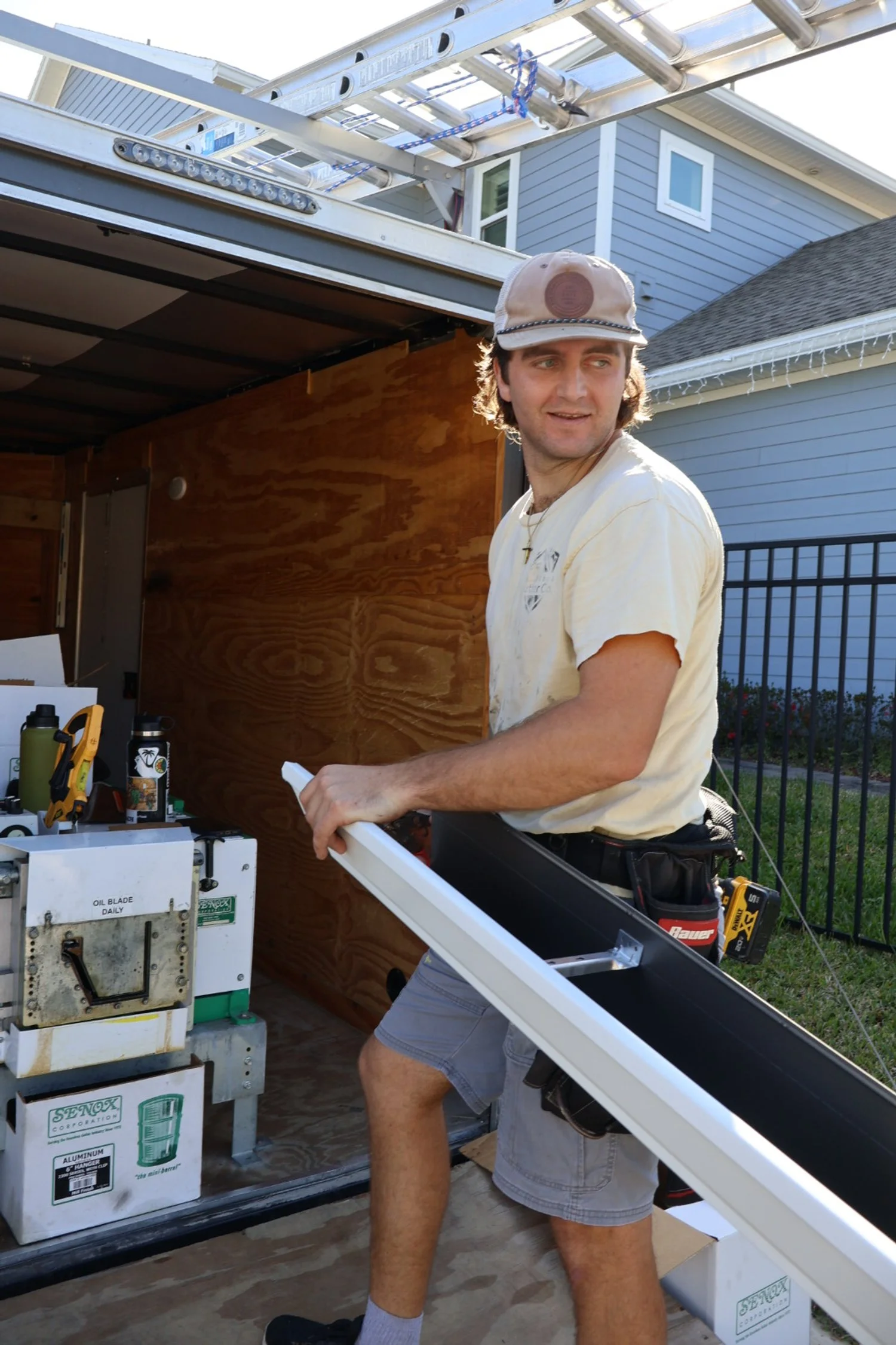 A man wearing a beige cap and white T-shirt is holding an outdoor window frame, standing next to a mobile workshop or storage trailer with tools and equipment inside on a sunny day.