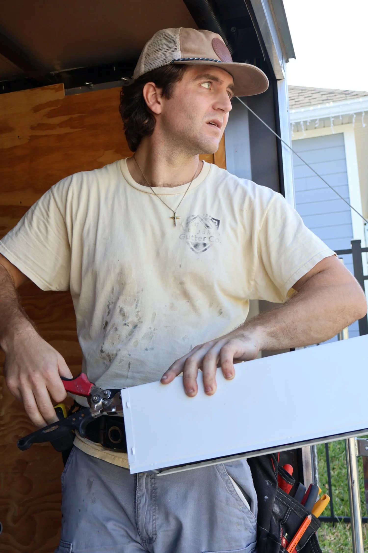 A man wearing a beige cap and a light-colored T-shirt is working on a white electrical panel or box, using a pair of pliers. The man has a cross necklace around his neck and is standing in a partially enclosed outdoor space.
