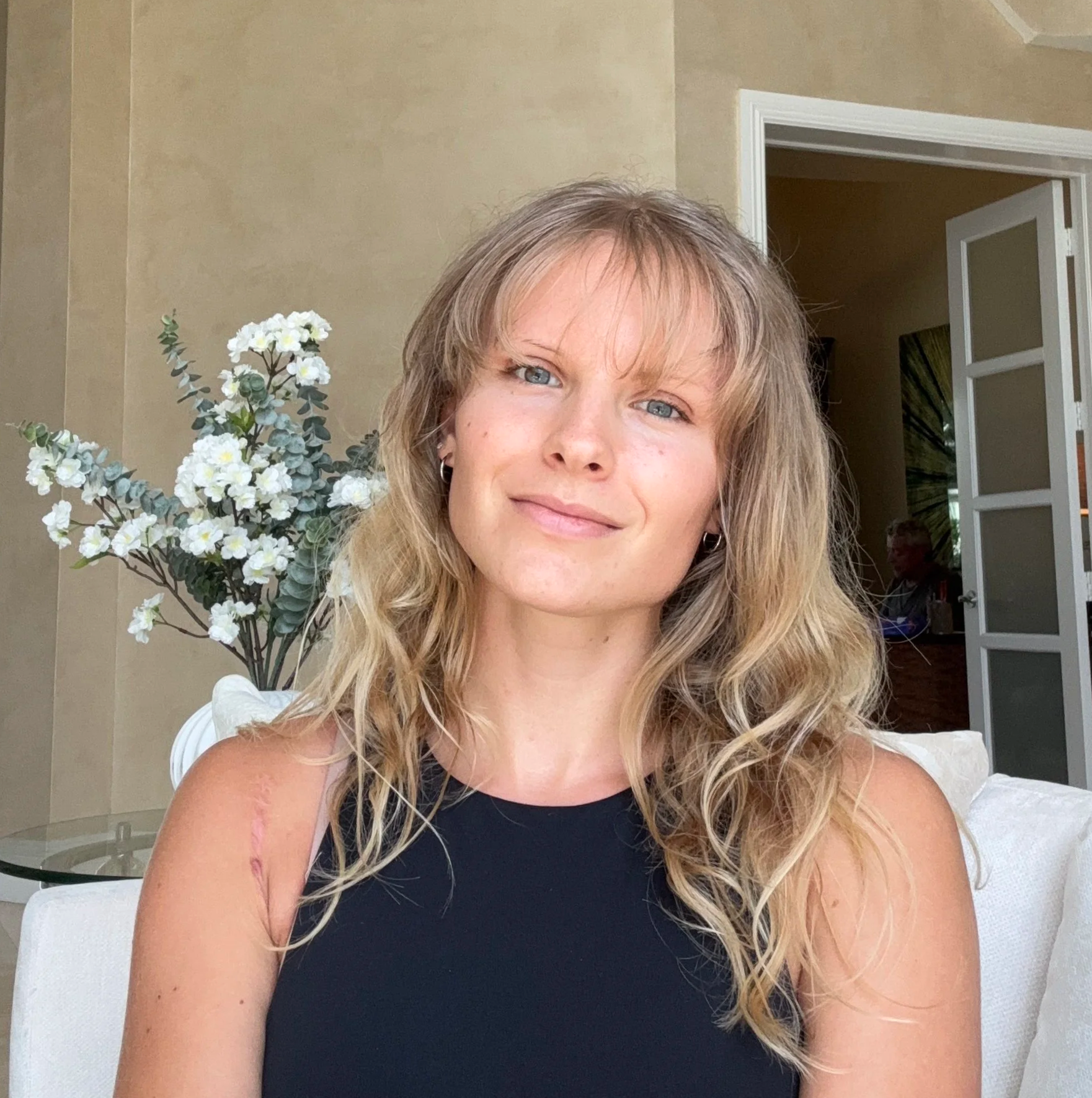 Close-up of a smiling woman with wavy blond hair, wearing a black sleeveless top, sitting indoors with a floral arrangement and open door in the background.