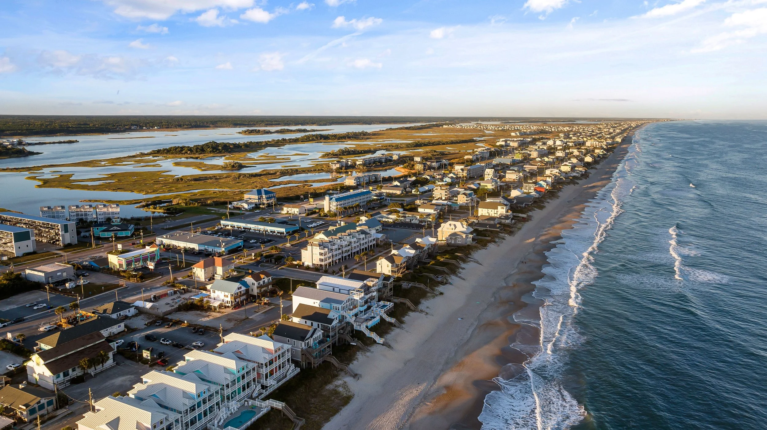 Aerial view of a coastal city with high-rise buildings along a sandy beach, ocean, and waterways with boats and marshlands.