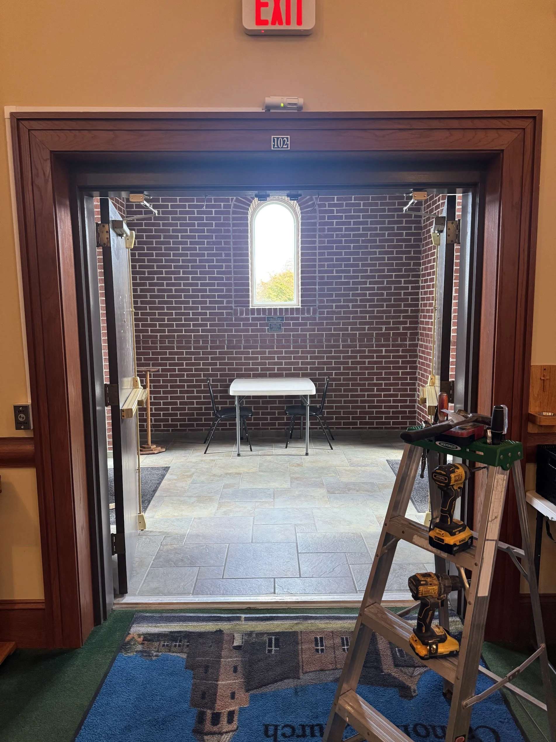 An incomplete interior door installation with tools on a step ladder, a brick wall with a window in the background, and a room number 102 sign above the door frame.