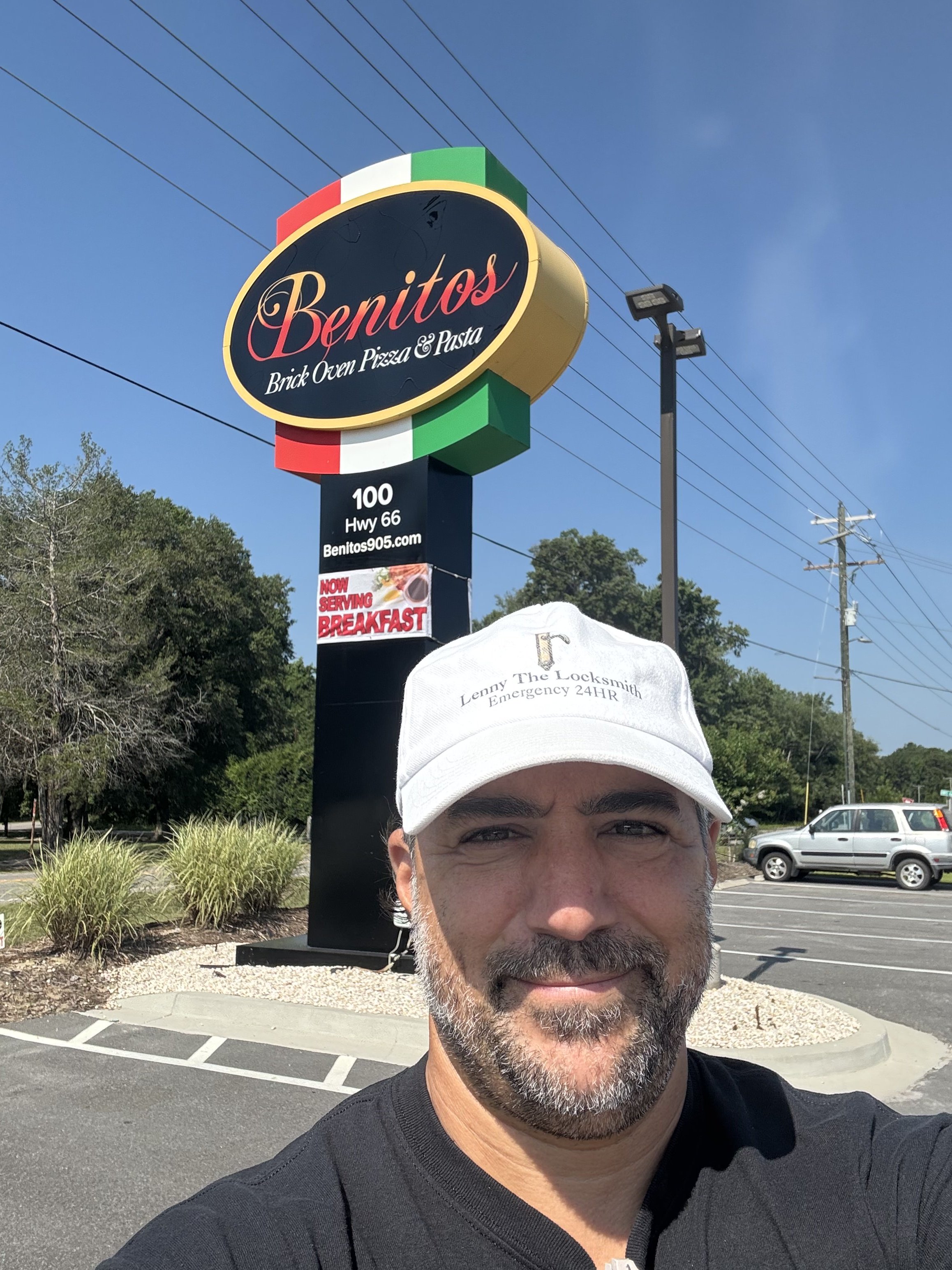 A man with gray hair and beard, wearing a white cap and black shirt, taking a selfie outdoors in a parking lot with trees, parked cars, and a sign for Benito's Brick Oven Pizza & Pasta in the background.