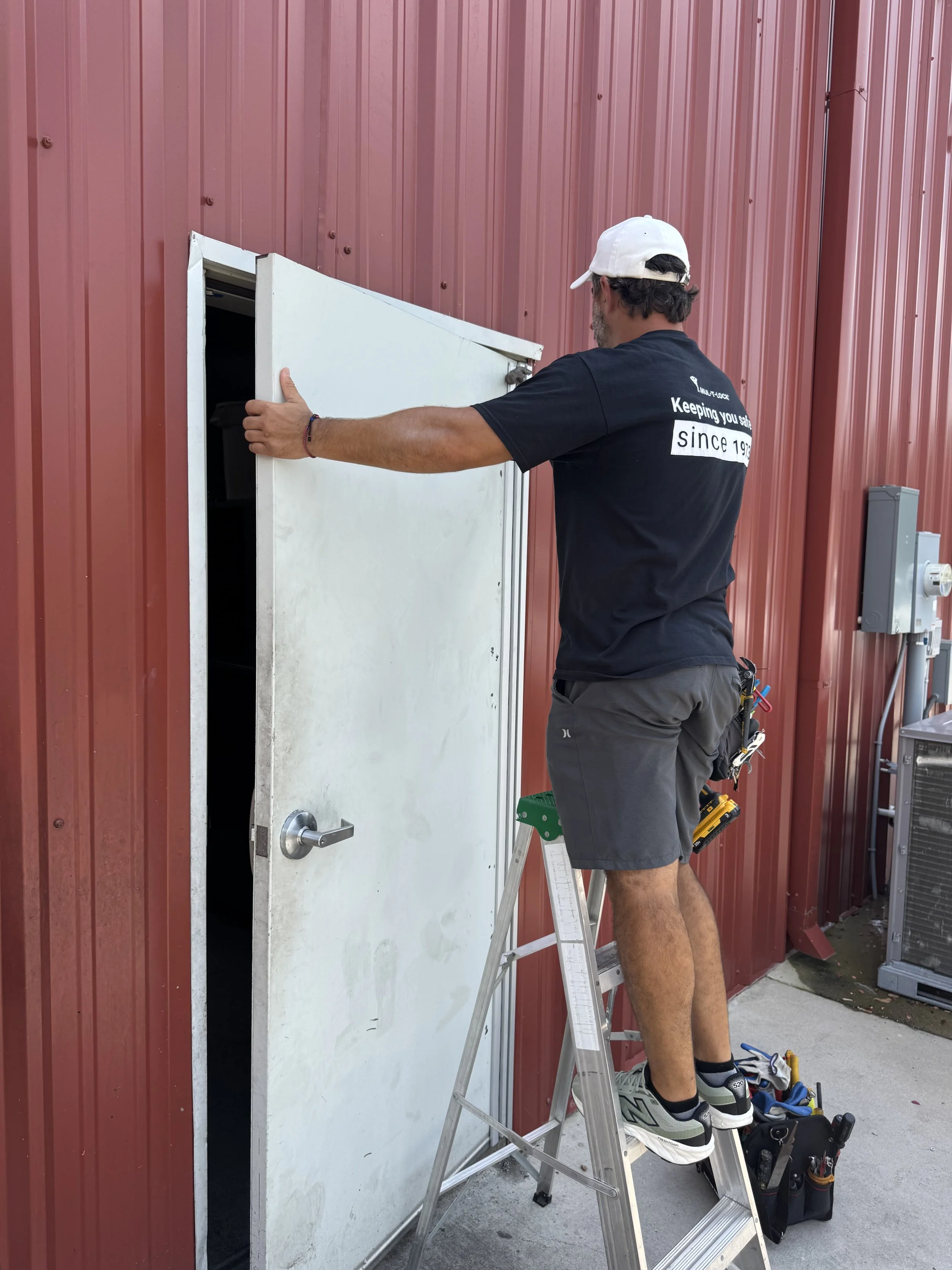 A man wearing a black T-shirt, gray shorts, and gray sneakers is standing on a ladder in front of a white door with a small window, working on the door handle. He has a white cap and a tool belt with various tools hanging on his right side. The background features a red metal building wall, and there are electrical panels and equipment nearby.