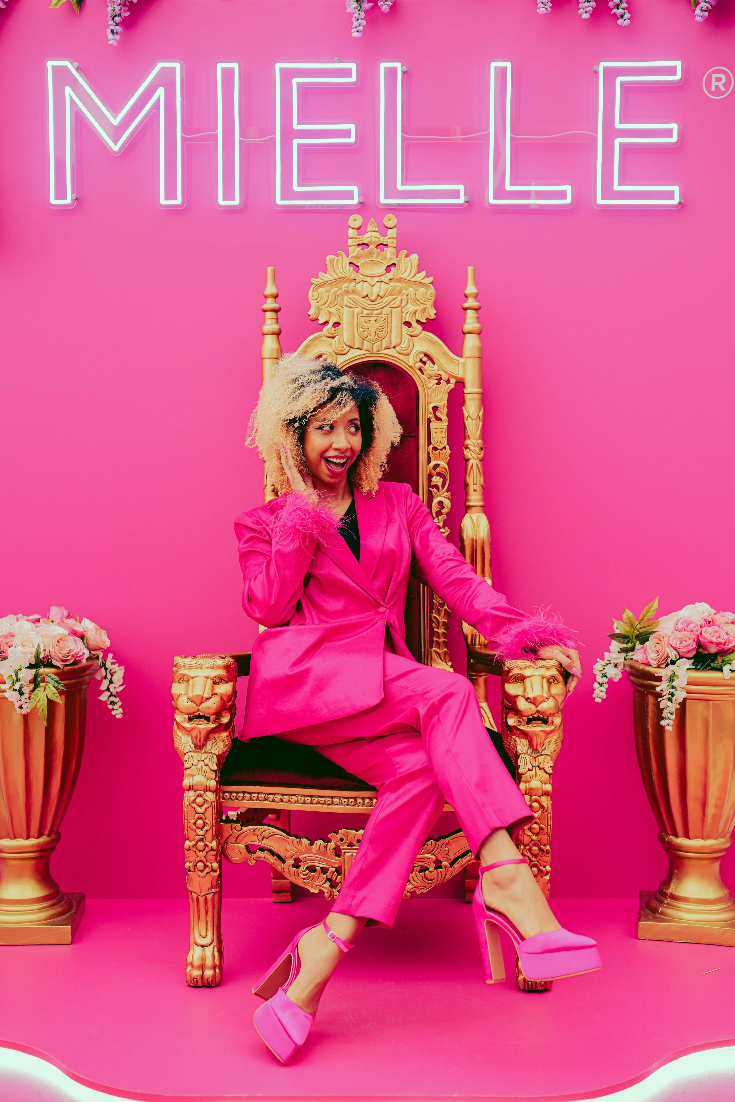 A woman with curly blonde hair, dressed in a pink suit and high heels, sitting on an ornate golden throne on a pink background. Behind her, a neon sign reads 'MHELLE'. Flowers in gold vases flank her on both sides.