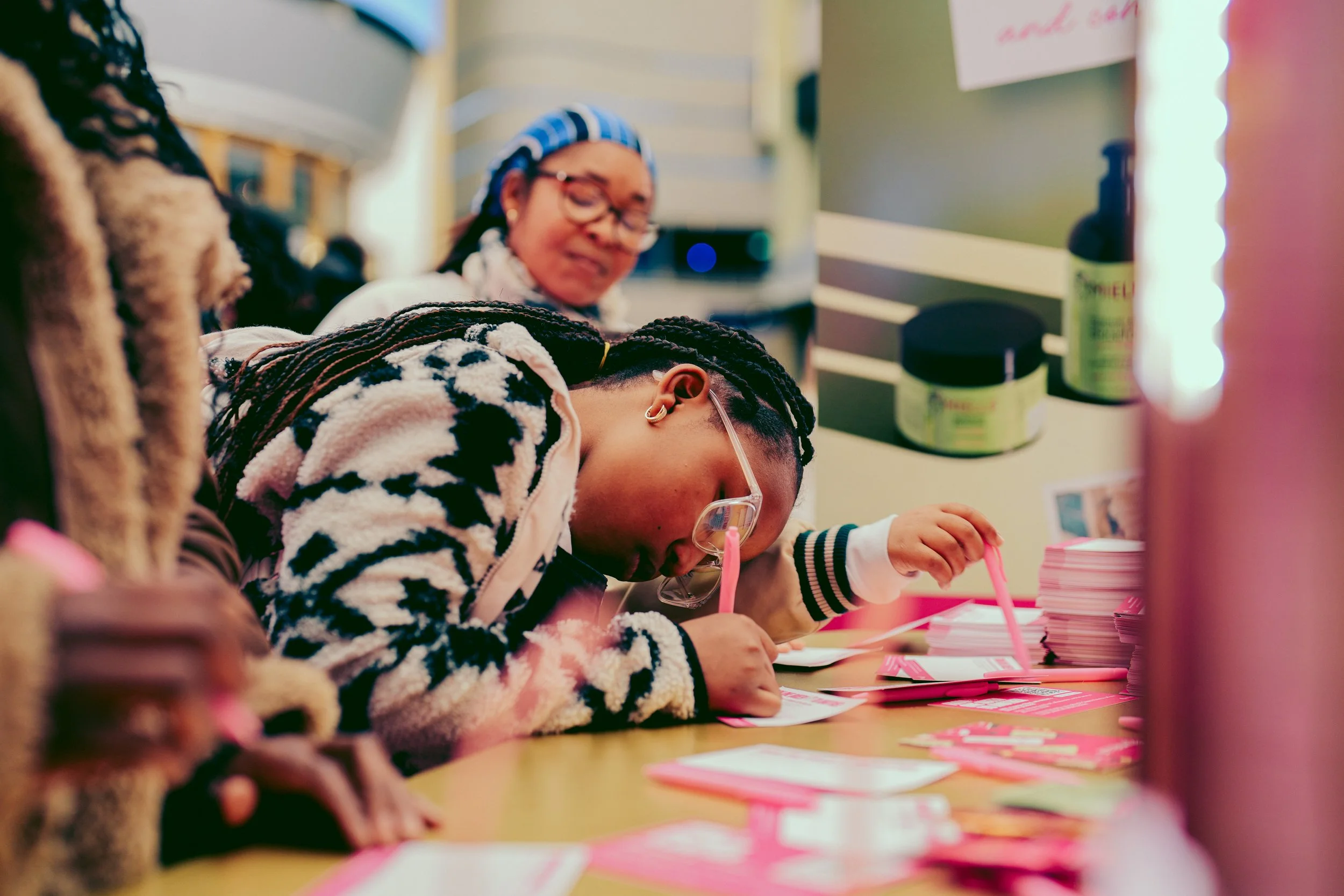 Young girl with braided hair and glasses writing with a pink pen at a table covered with pink papers, with an older woman standing behind her.