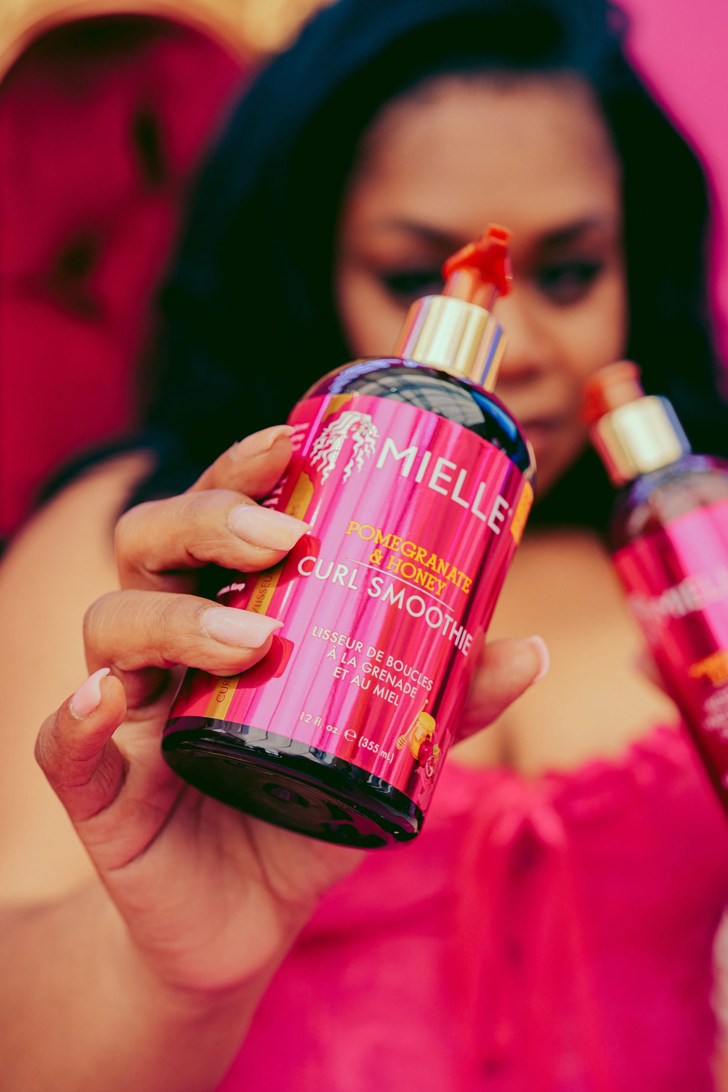 A woman holding a bottle of Mielle Pomegranate & Honey Curl Smoothie hair product, with another similar bottle in the background. The woman is wearing pink and has dark hair.