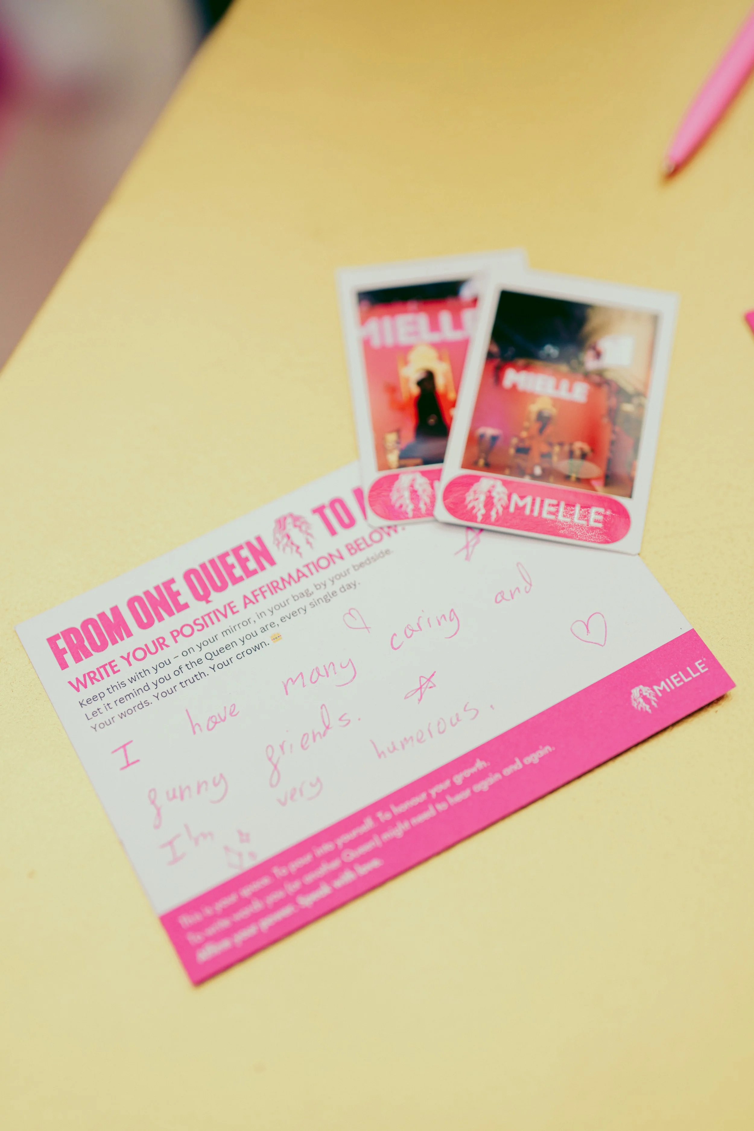 A pink and white affirmation card titled 'From One Queen To Another' with handwritten note and two instant camera photos on a yellow surface.