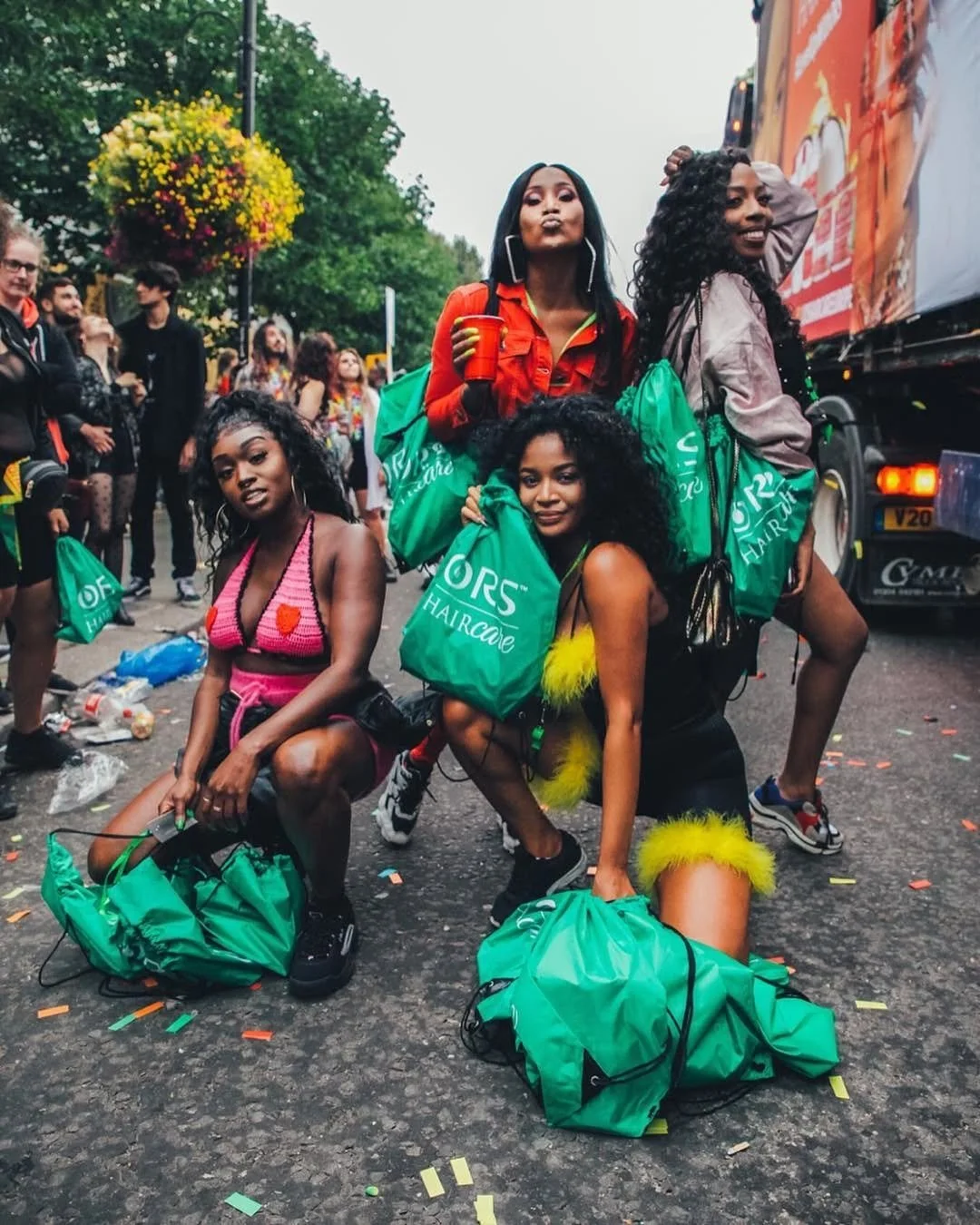 Four women wearing colorful outfits, with some holding green bags that say 'Ors Haircafe,' posing outdoors during a festival or parade, with a crowd and trees in the background.
