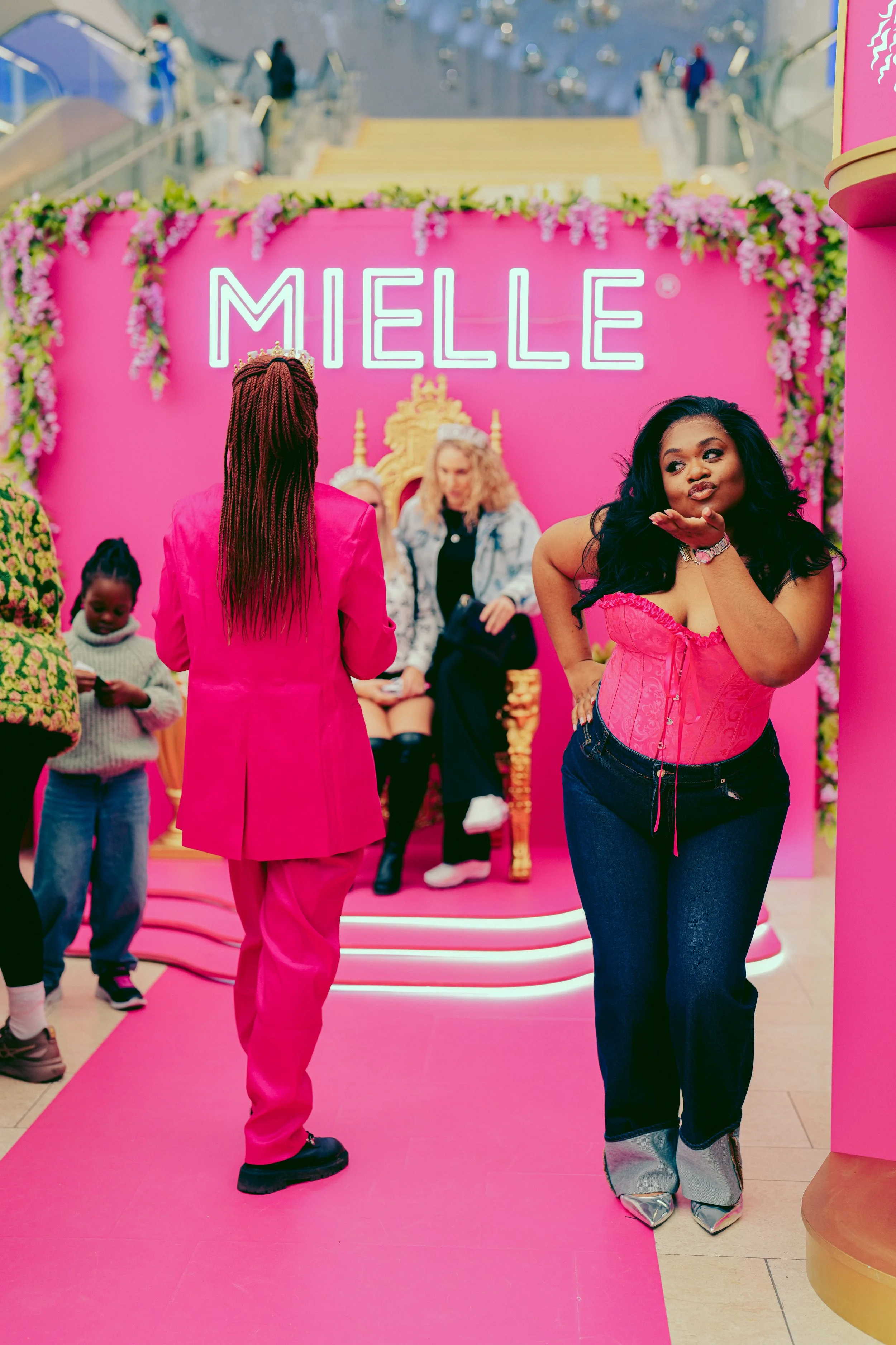A woman in a pink corset top and jeans posing in front of a pink backdrop with the name 'MIELLE' written in neon. The backdrop is decorated with pink flowers and a woman sitting on a golden throne is seated in the background.