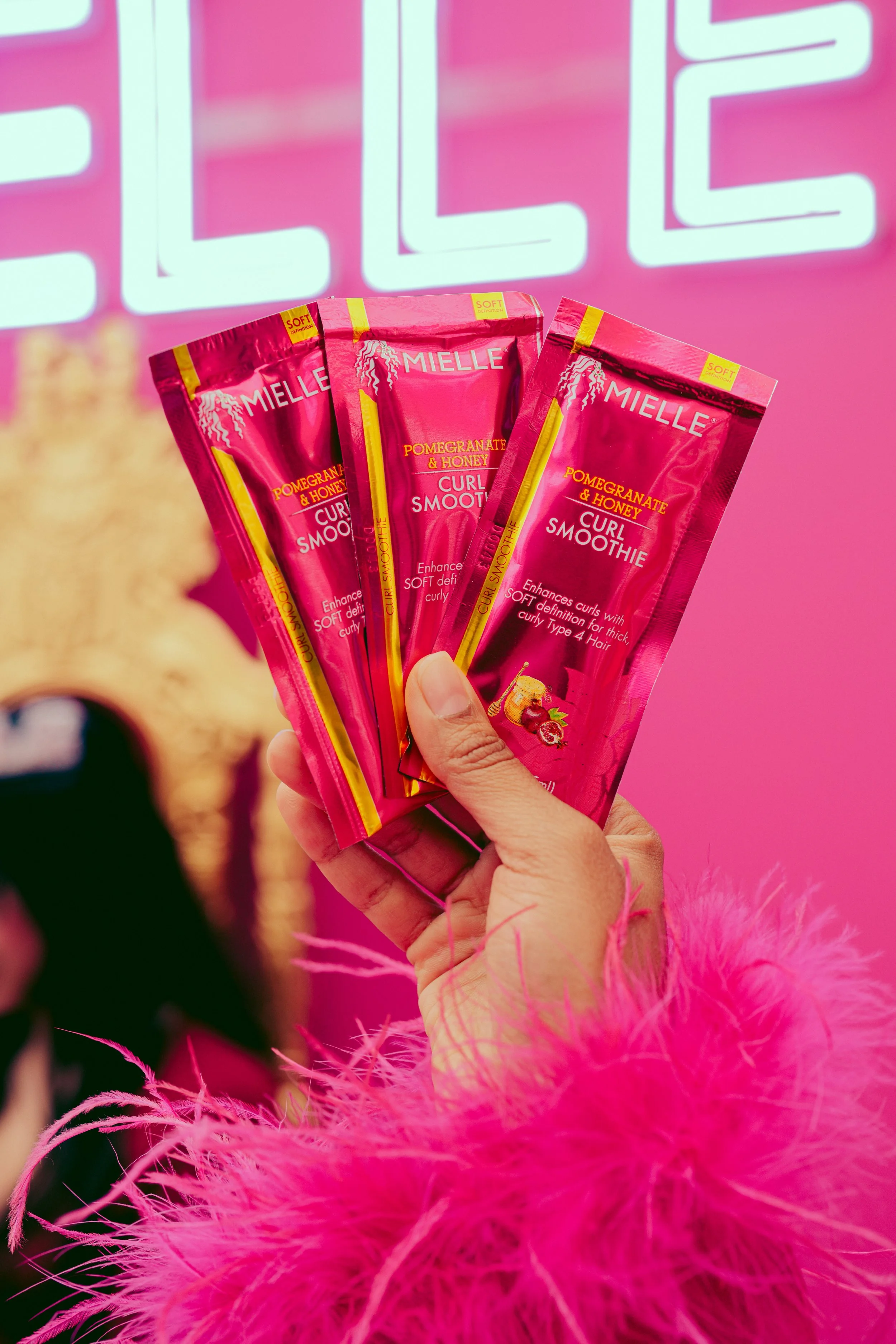Hand holding three packets of Mielle Pomegranate & Honey Curl Smoothie hair product against a pink background.