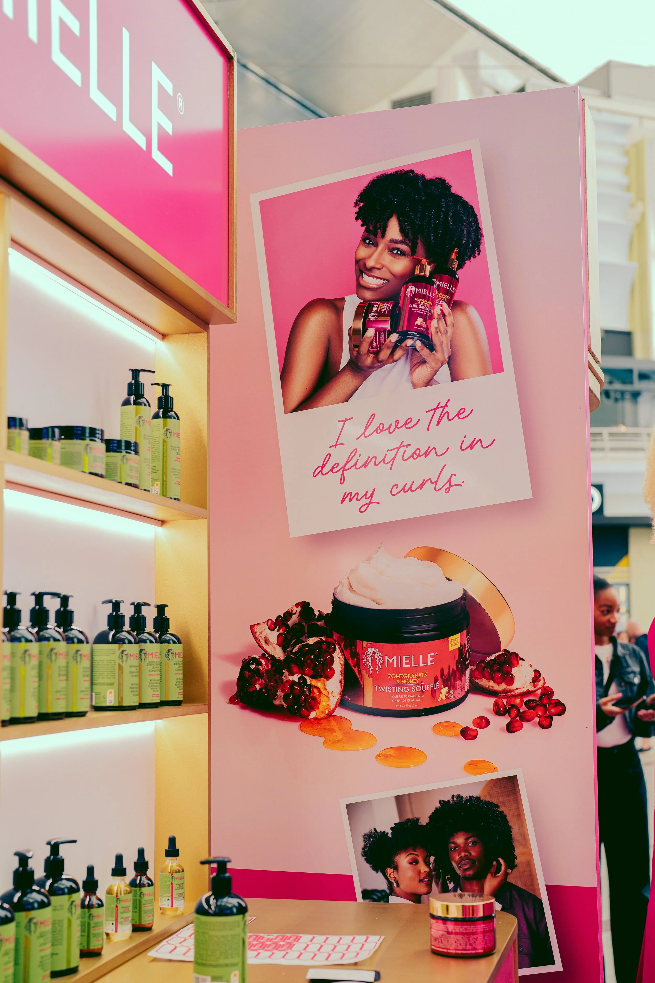 Beauty product display with a pink promotional poster featuring a woman with curly hair holding hair care bottles, the text online "I love the definition in my curls", with a jar of body cream and pomegranate imagery below, and various skincare produ