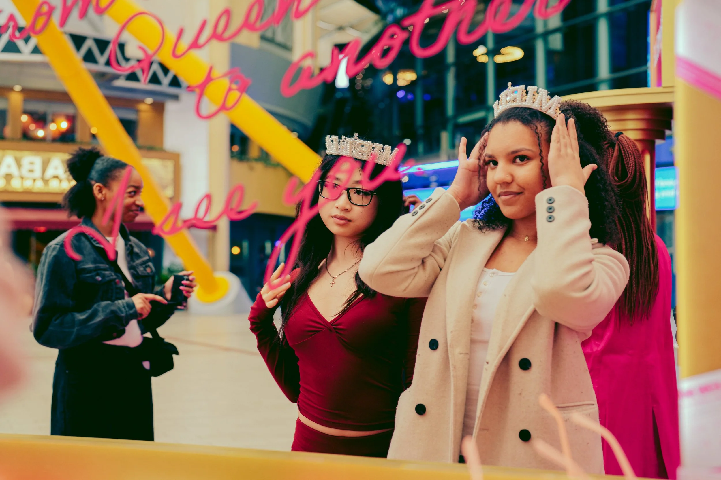Two young women trying on tiaras at an amusement park or carnival, with one adjusting her tiara and the other holding her head. In the background, another woman in a denim jacket is looking at her phone, and colorful amusement park rides and signs ar