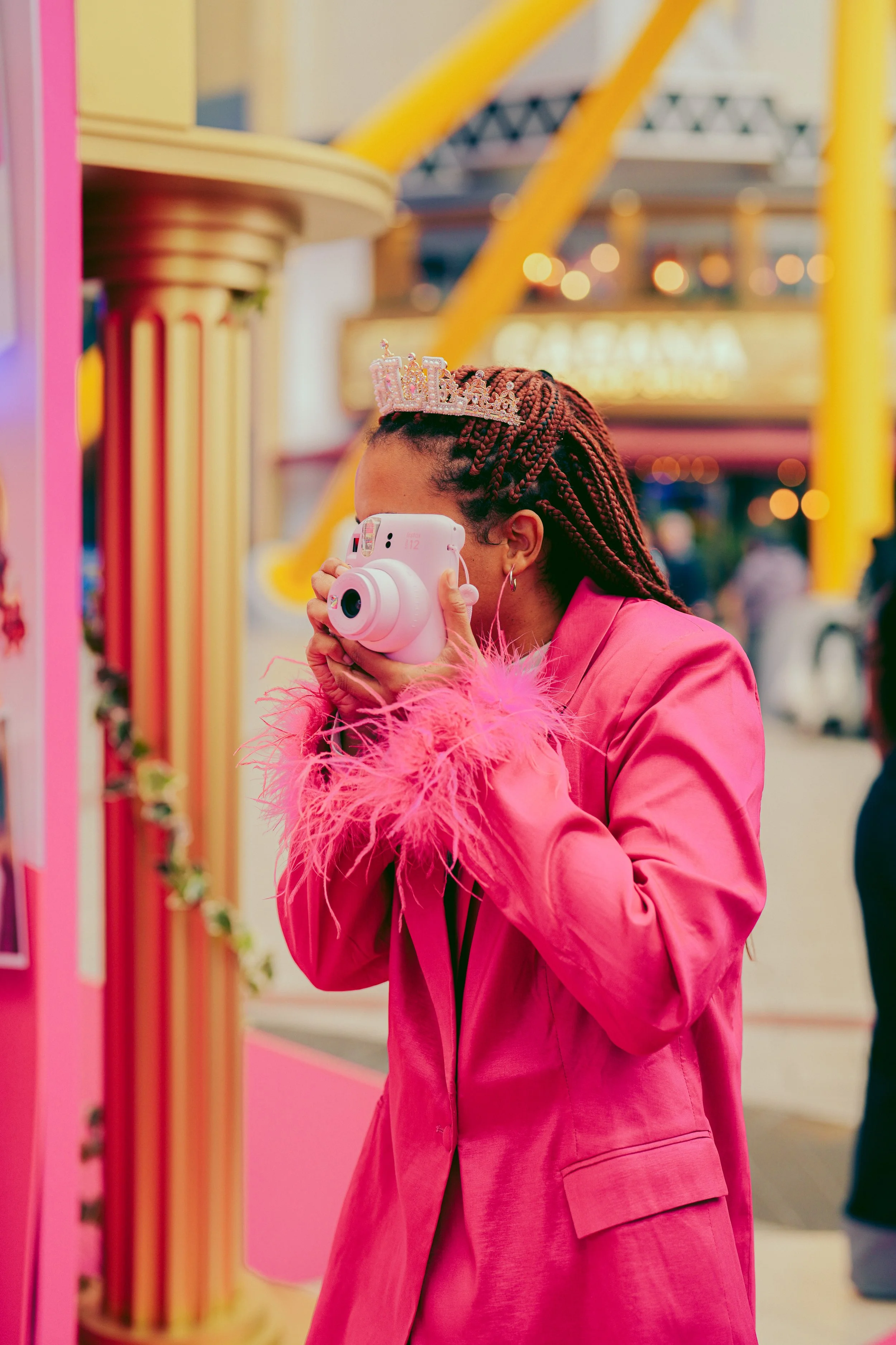 A woman with braided hair wearing a pink jacket and a tiara taking a photo with a pink instant camera at an amusement park or fair.