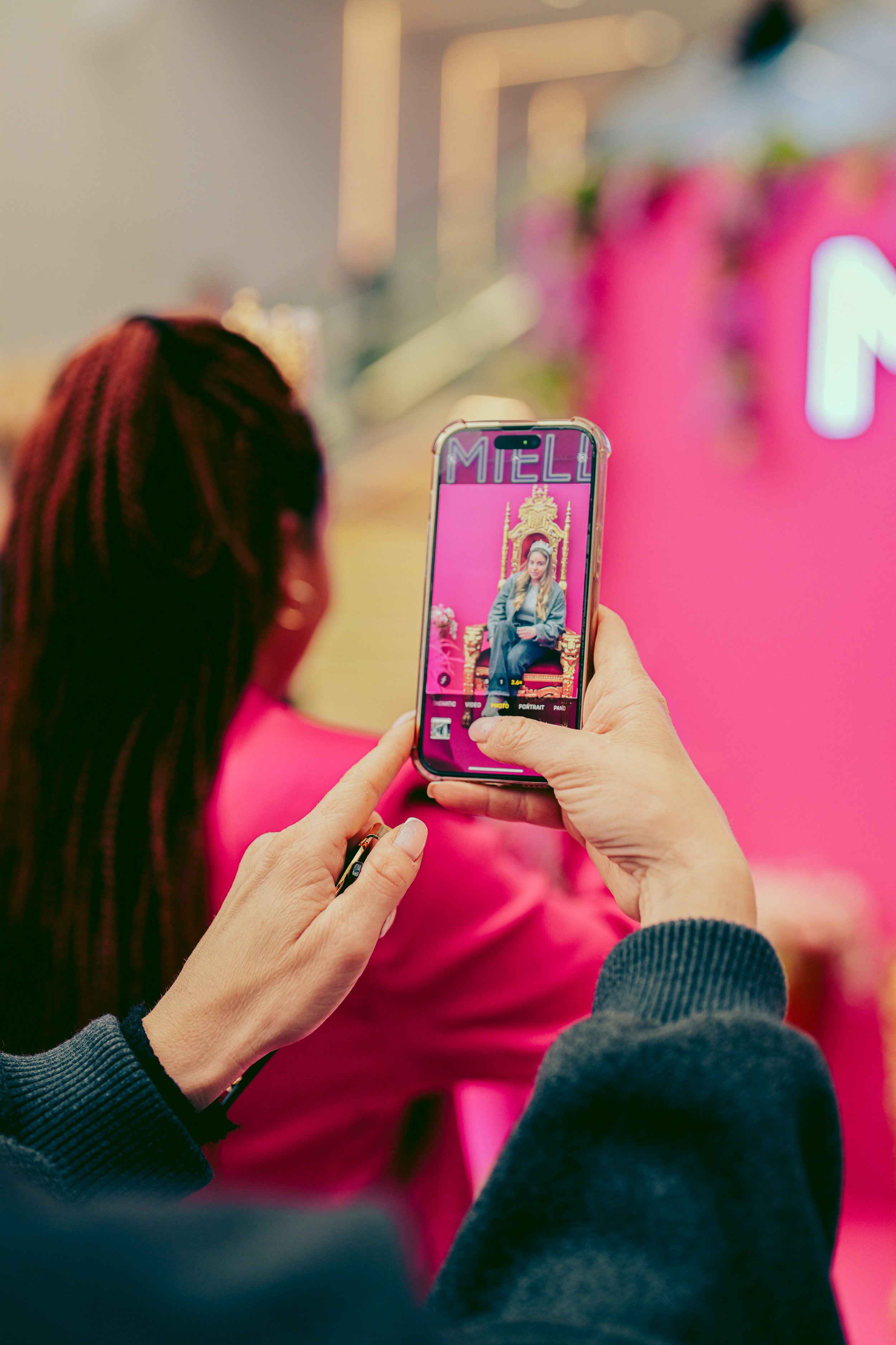 A person taking a photo of a woman sitting on a throne with a pink background using a smartphone, with pink and gold decor around.