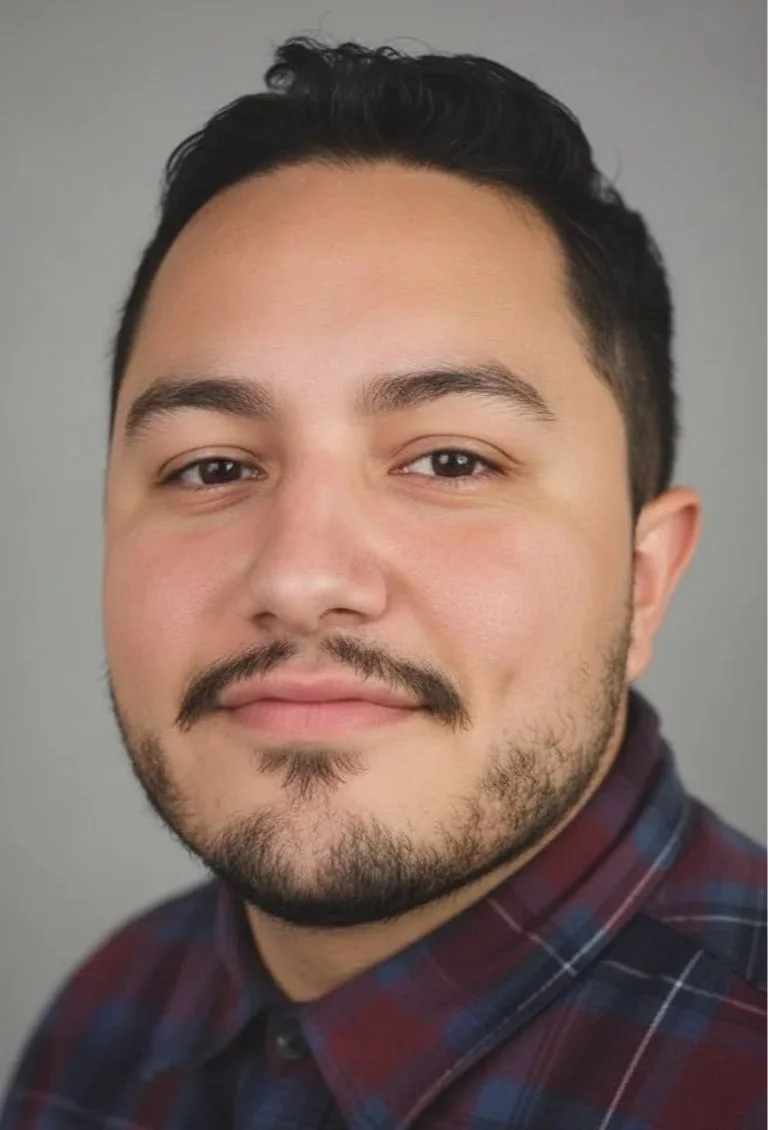 A close-up portrait of a young man with dark hair, facial hair, and light skin, wearing a plaid shirt, against a plain gray background.