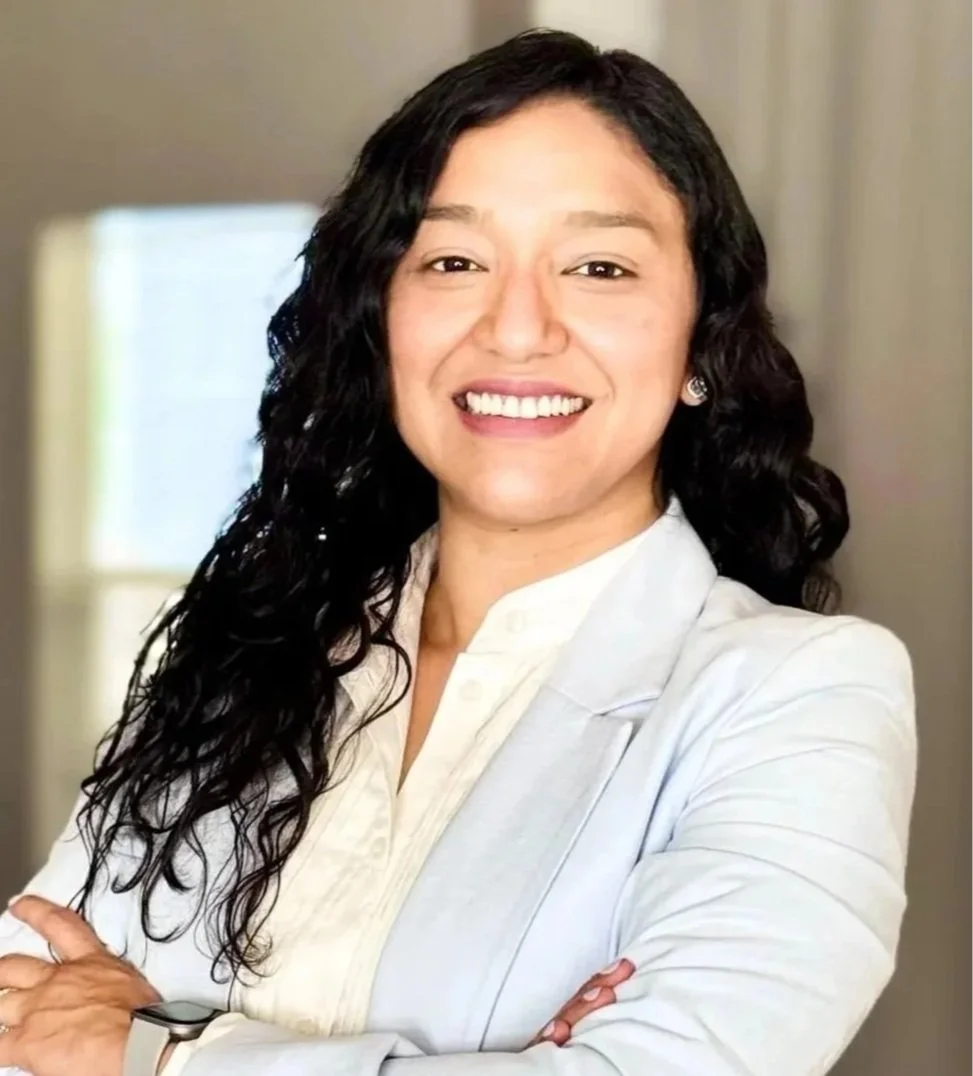 Professional woman with long, curly black hair smiling, wearing a white blazer and earrings, standing with arms crossed inside a well-lit room.