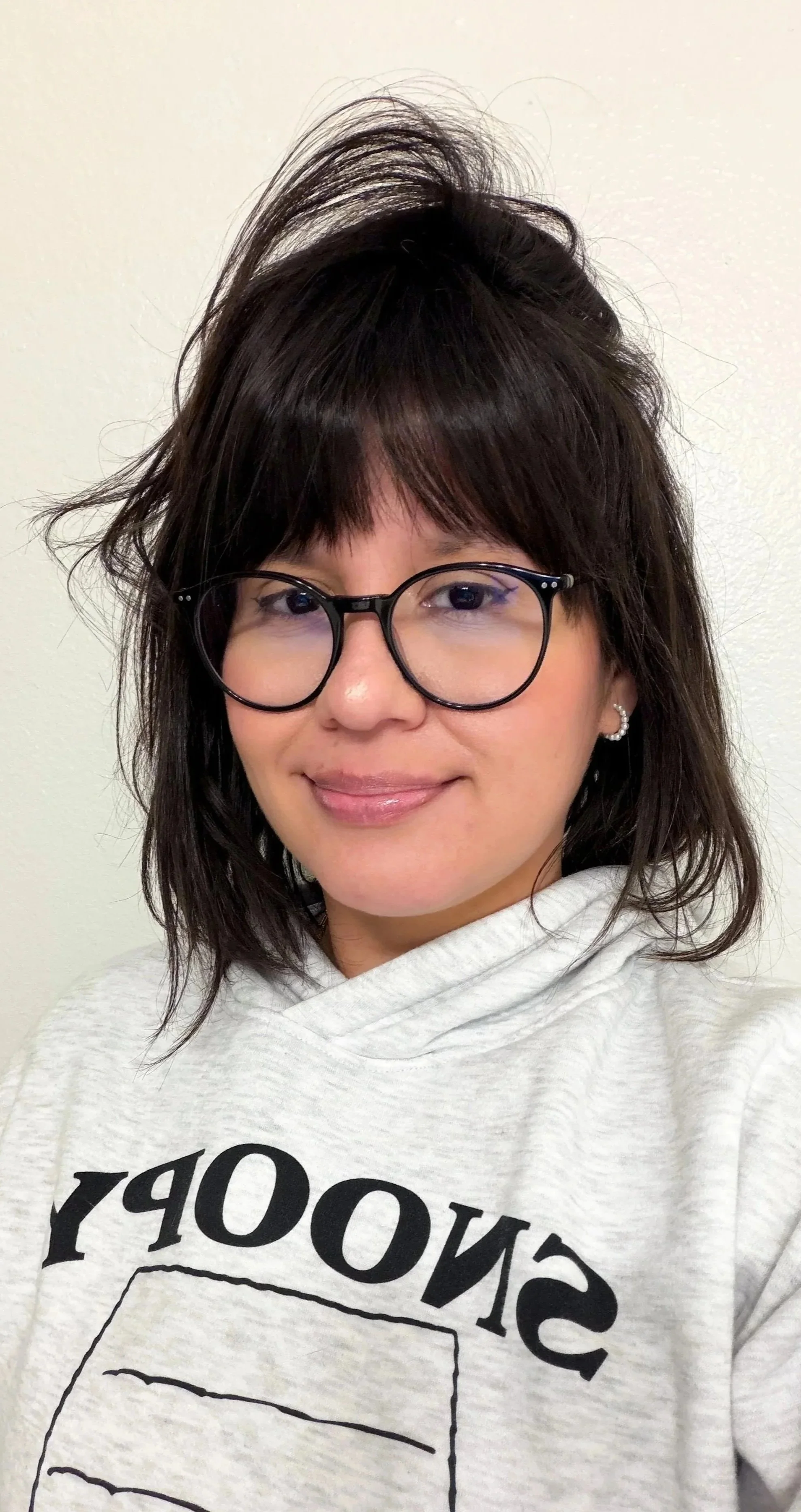 A woman with shoulder-length dark hair, glasses, and earrings, smiling at the camera, wearing a white hoodie with black text that says 'SNOOPY' and a graphic of a lined notepad.