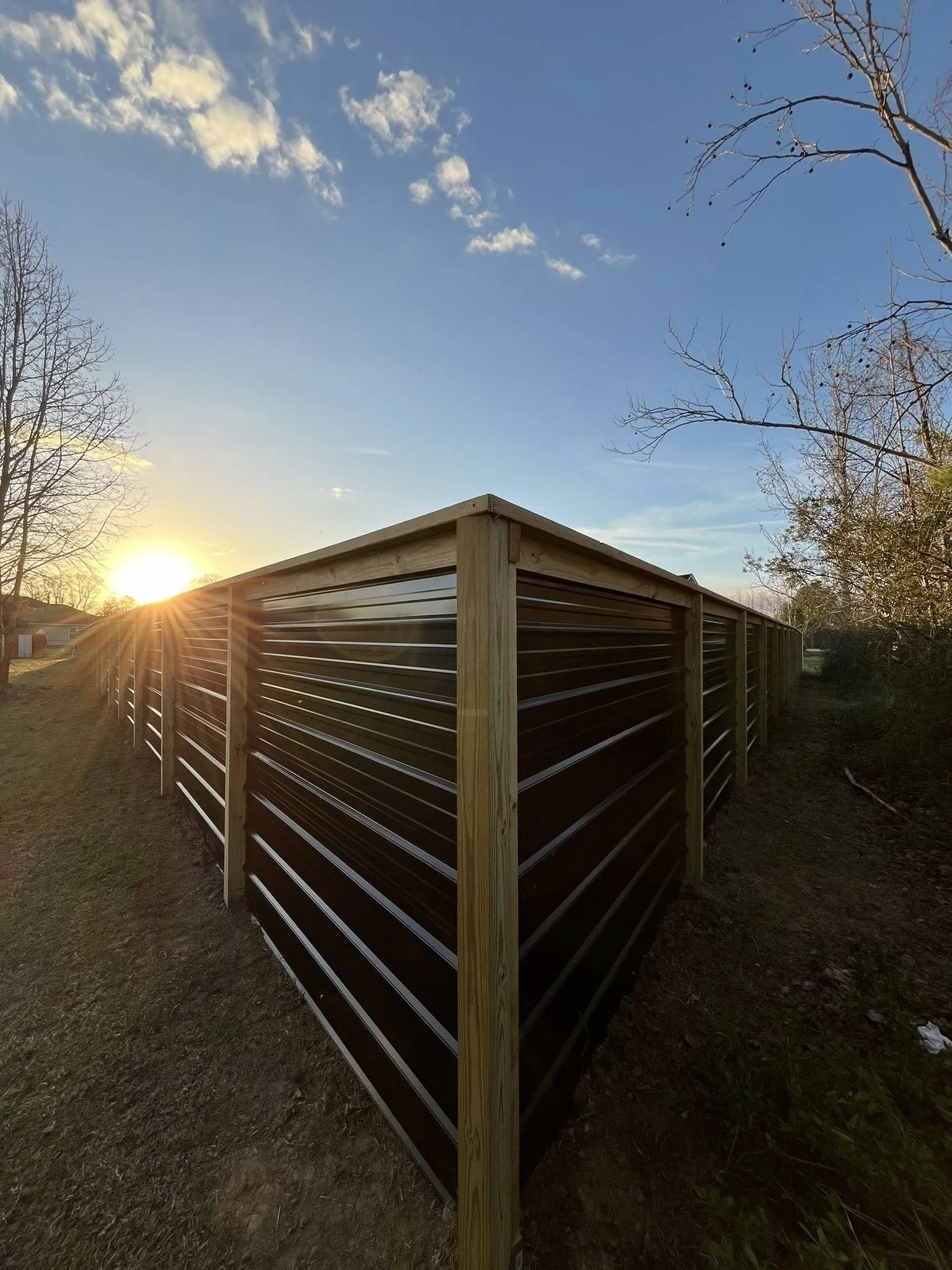 Sunset over a dark brown metal fence with wooden posts, set on grass, with trees and a blue sky with clouds in the background.