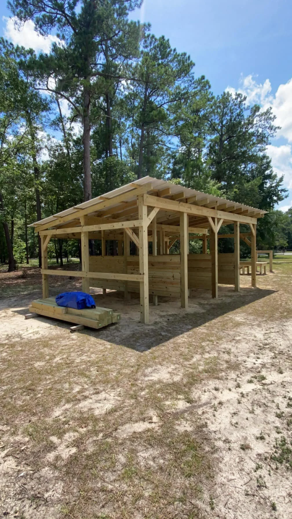 A wooden structure is under construction in a park, surrounded by tall trees and a partly cloudy sky.