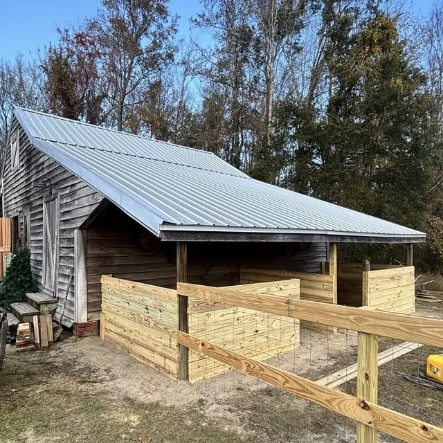 A wooden stable built around a barn with a metal roof, located outdoors with trees in the background.