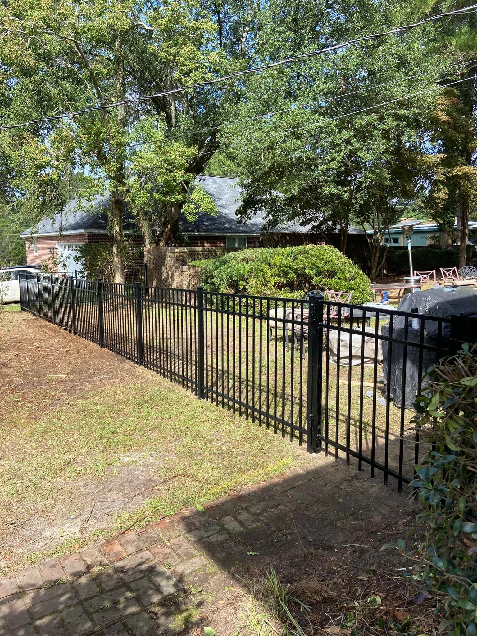 Black custom metal fenced backyard with trees, bushes, and outdoor furniture, adjacent to a brick house with a garage in Sumter, SC