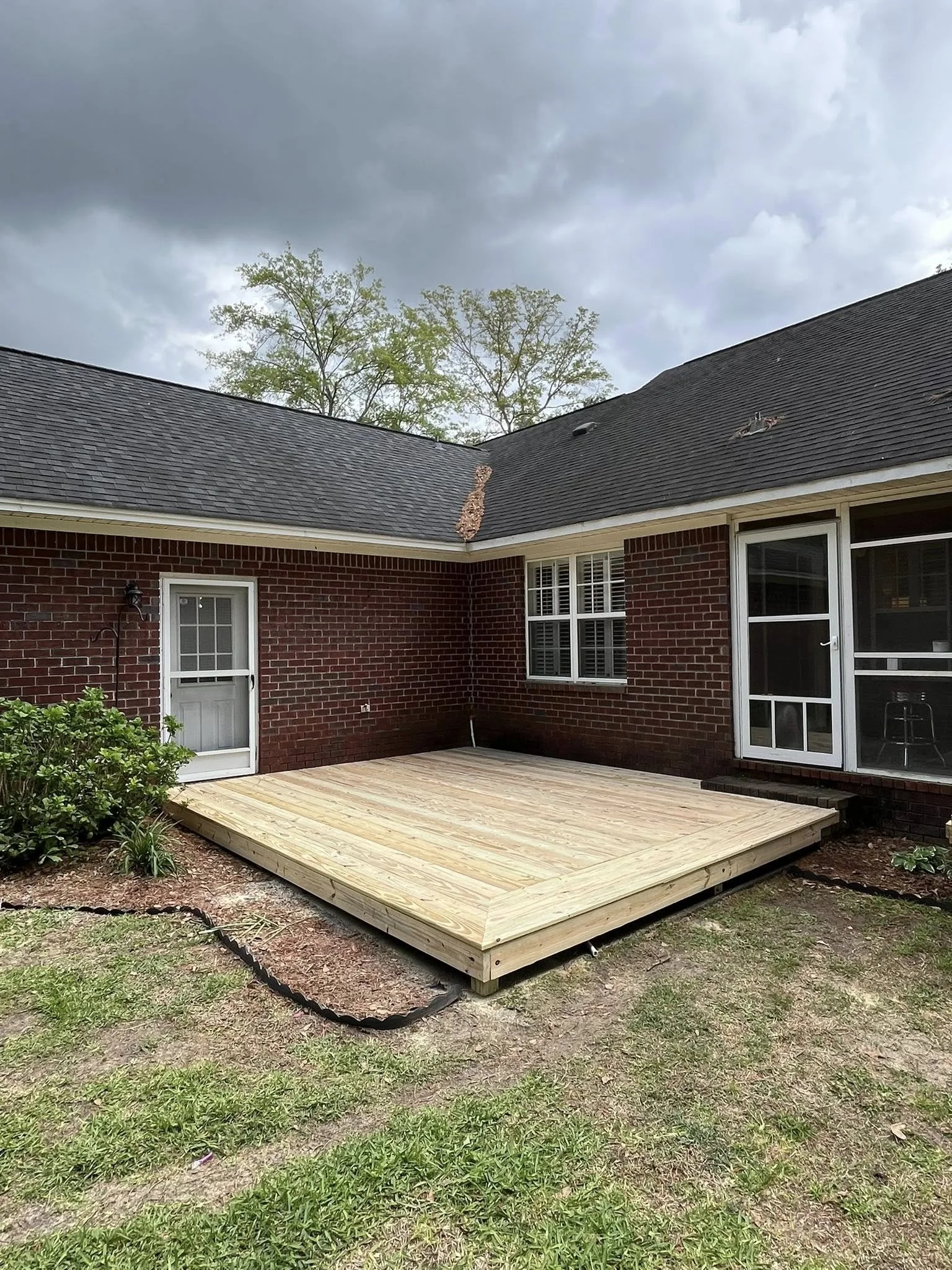 A newly built wooden deck outside a brick house with a sliding door, a window, and a small door, under dark stormy clouds.