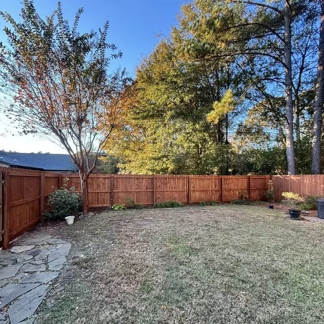 A Sumter backyard with a grass lawn, a wooden fence, built and stained by J Co., and several trees with autumn leaves. There are some potted plants along the fence and a stone patio area on the left.