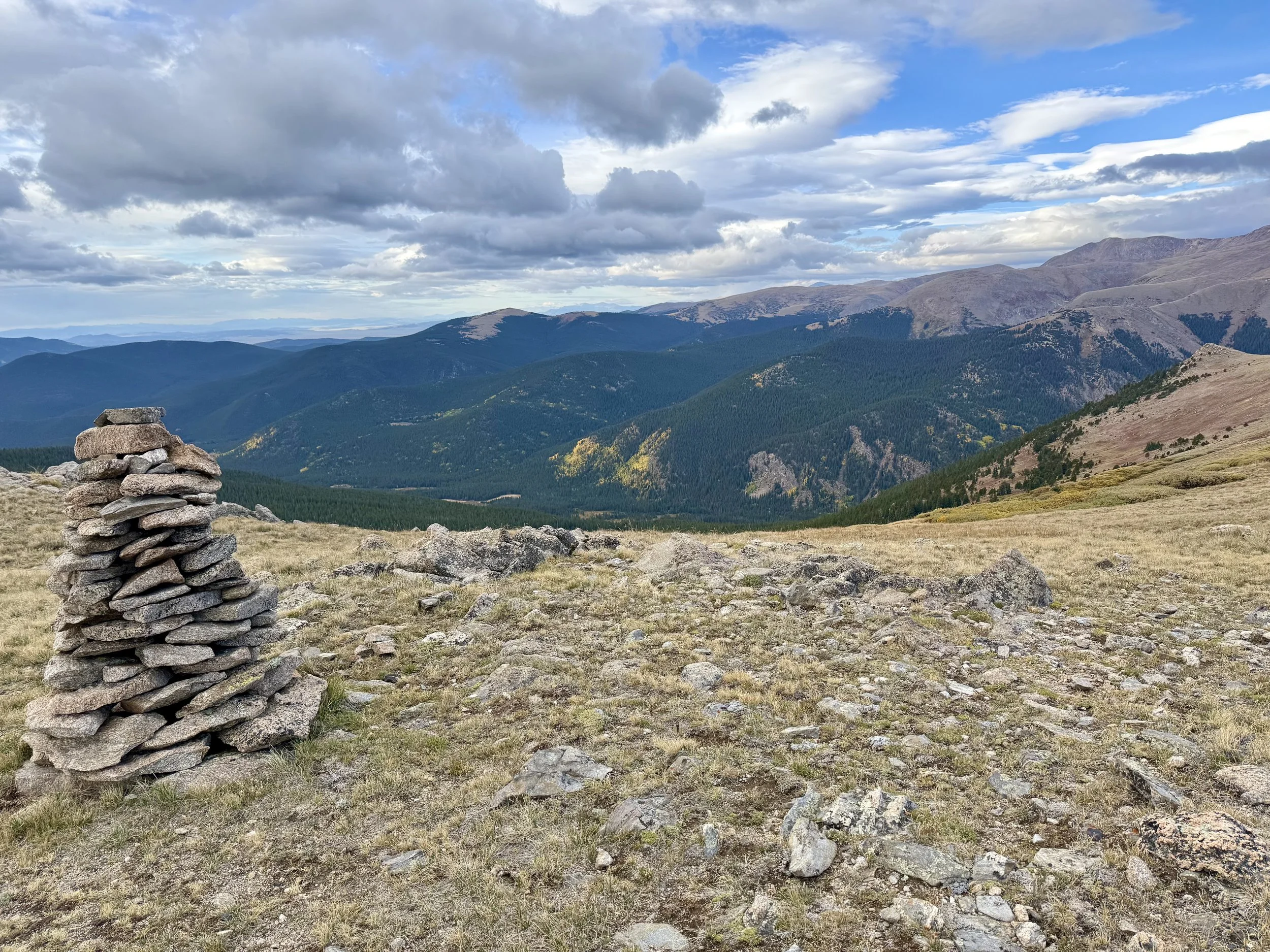 Stacked stone cairn on a mountain ridge overlooking forested hills under a cloudy sky.