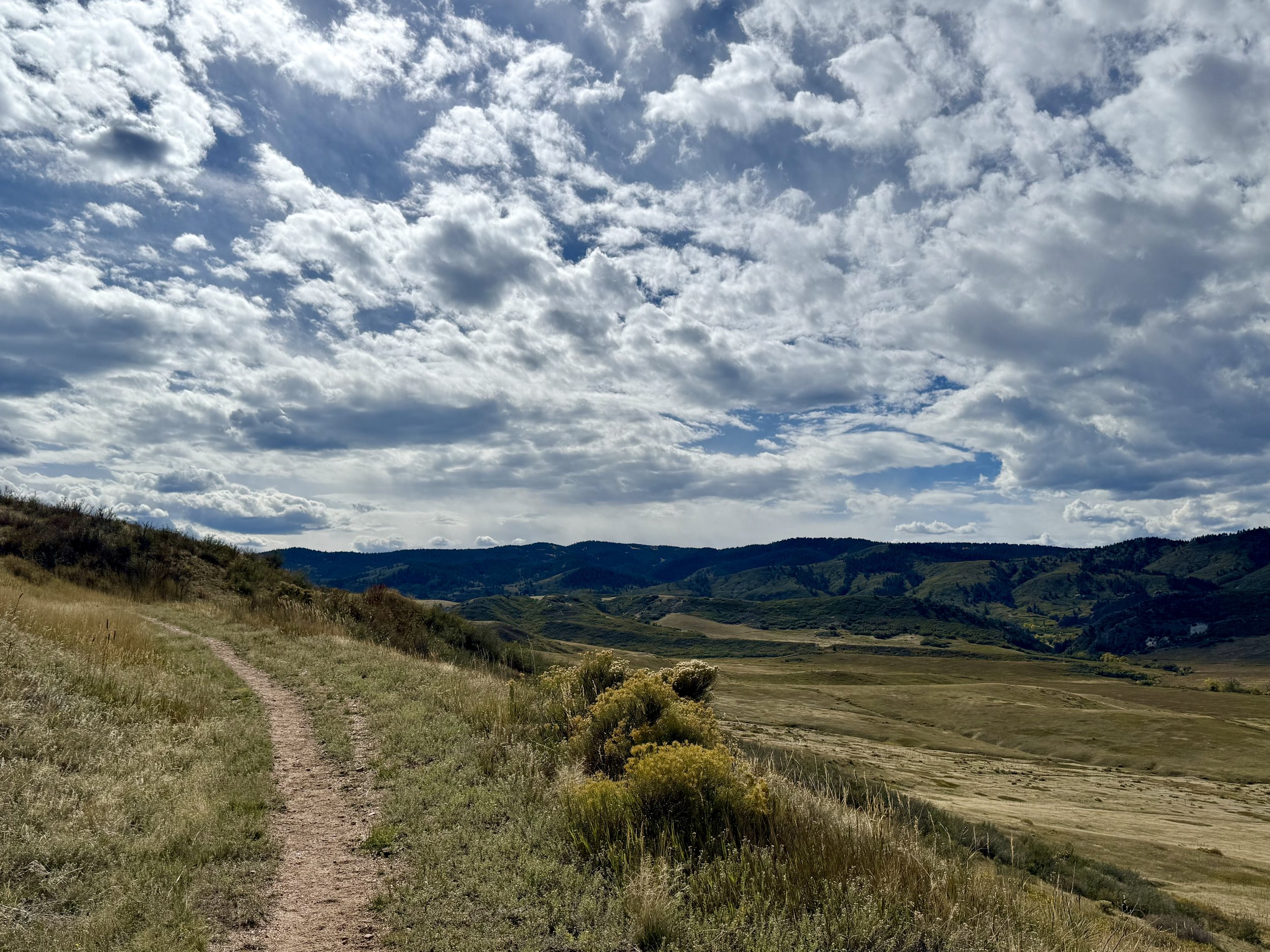 A dirt trail running through grassy hills under a partly cloudy sky with mountains in the distance.