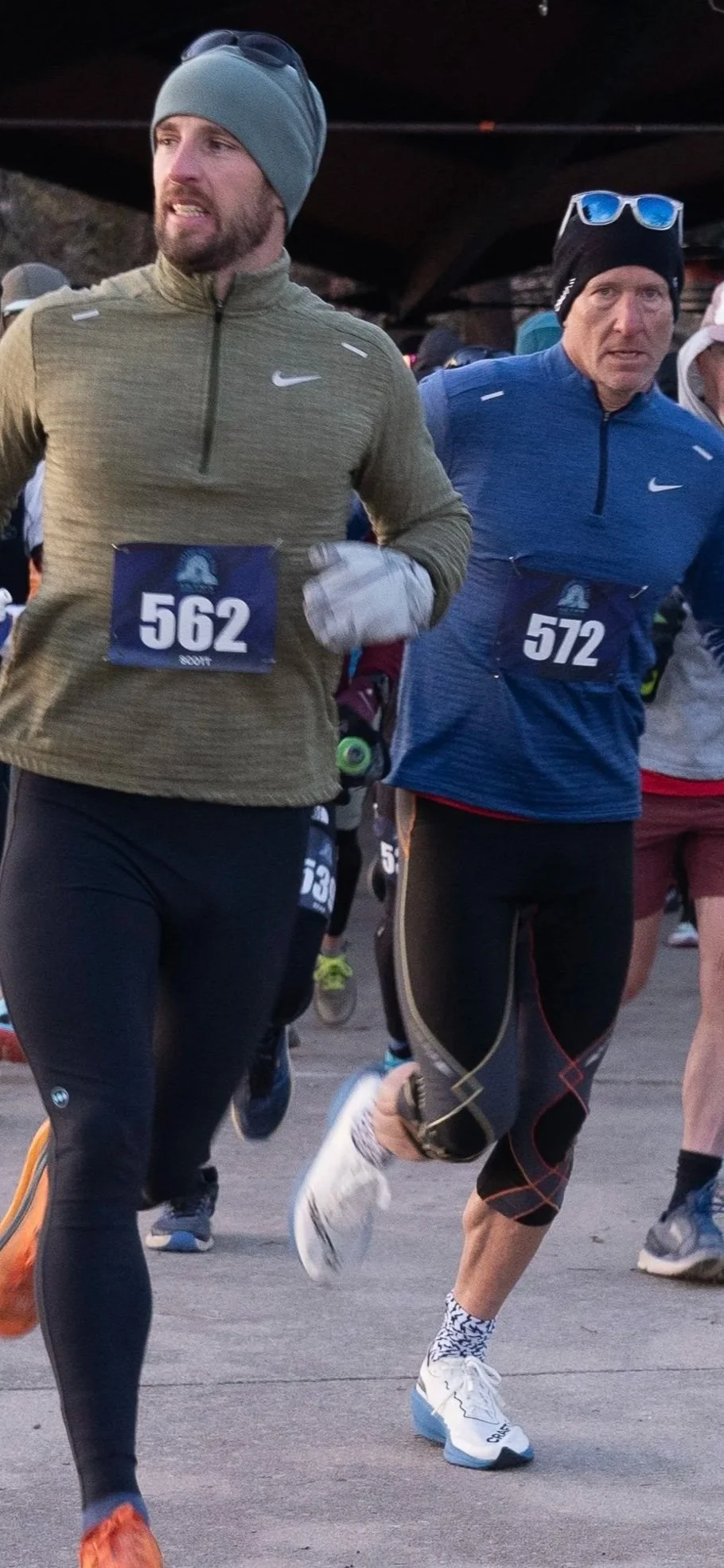 Two men participating in a marathon race, wearing bib numbers 562 and 572, running outdoors in athletic gear including jackets, gloves, and hats.