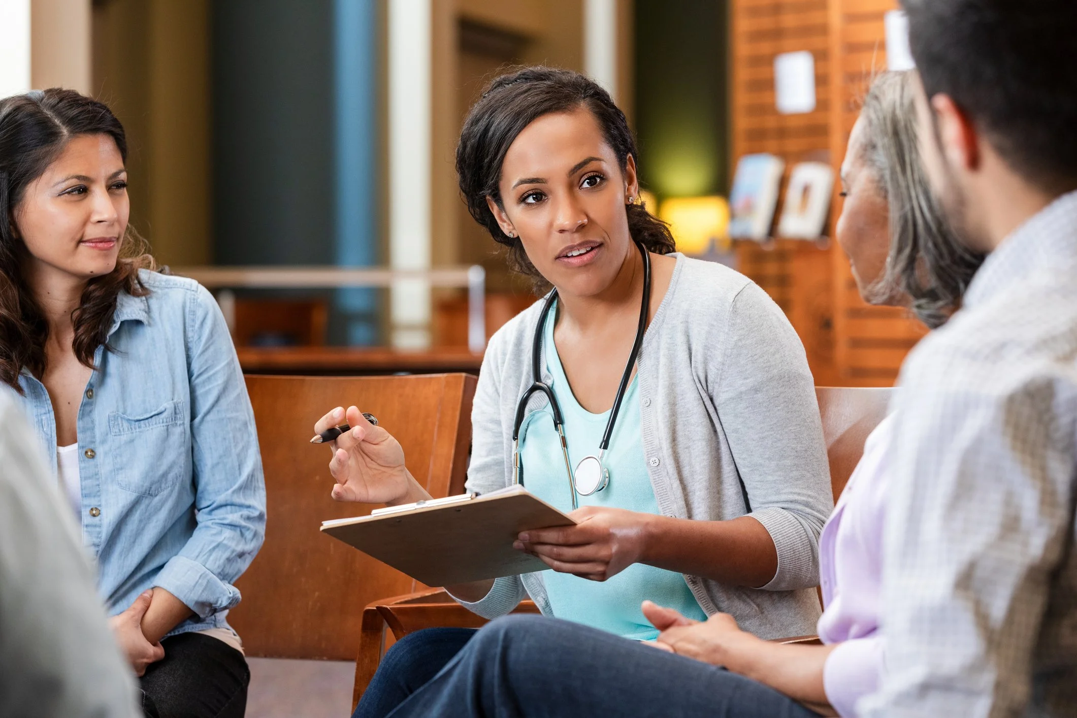 A female healthcare professional with a stethoscope around her neck talks to a group of diverse women in a waiting room or consultation area.