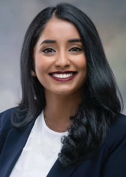 A smiling woman with long black hair, wearing a navy blazer and white blouse, against a blurred background.