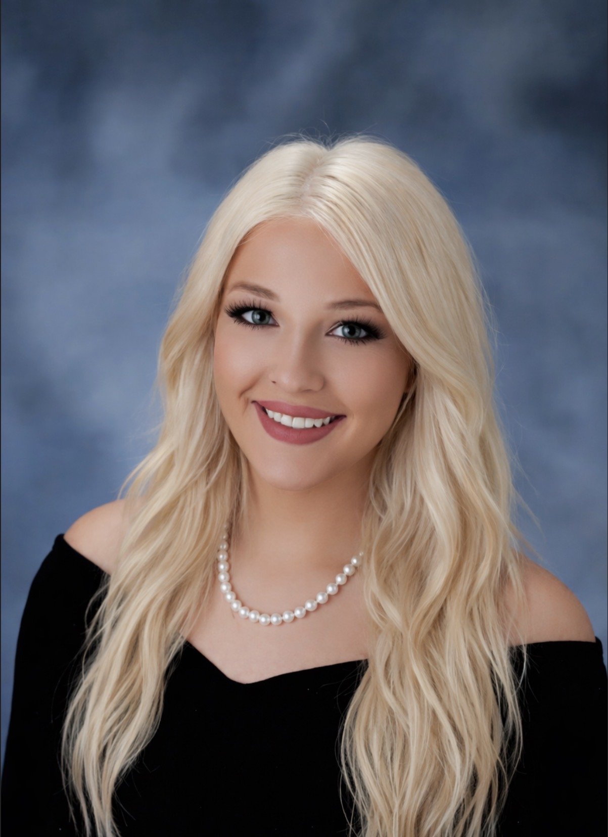 A young woman with long blonde hair, blue eyes, wearing a black off-shoulder top, a pearl necklace, and smiling against a blue background.