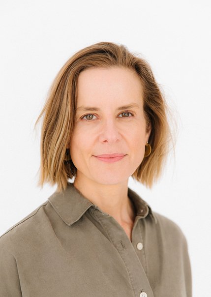 A woman with shoulder-length light brown hair, wearing a khaki shirt and small hoop earrings, standing against a white background.