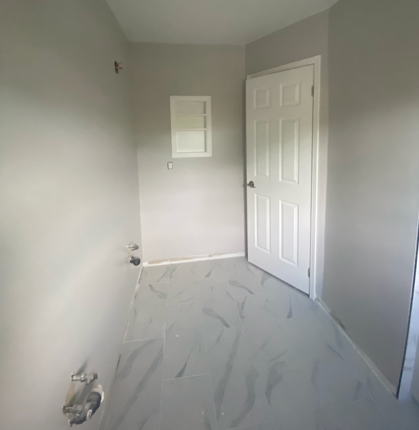 Empty laundry room with light gray walls, a white door, a small window, and plumbing fixtures for a washer and dryer, with gray and white marbled floor tiles.