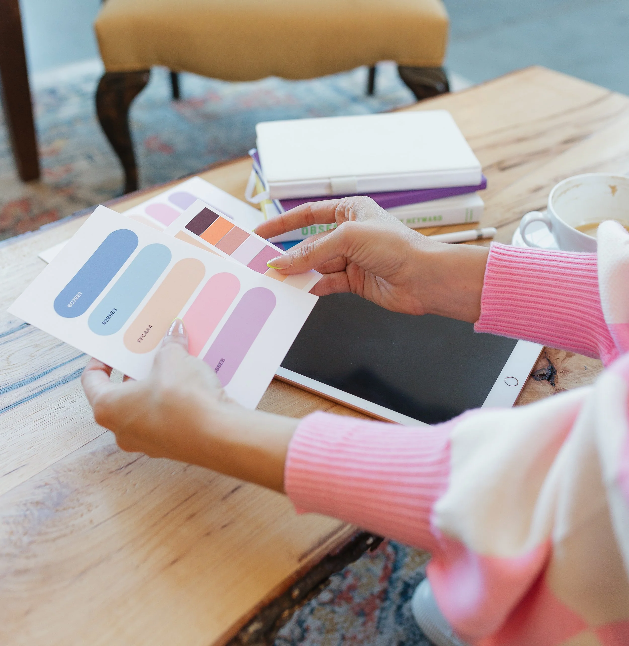 Person holding color swatches while sitting at a wooden table with a tablet, notebooks, and a coffee cup.