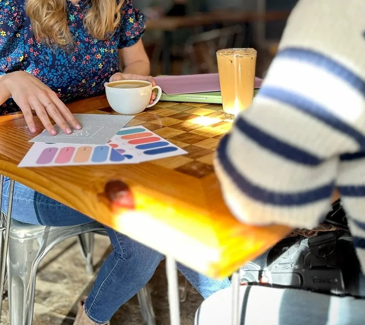 A woman at a wooden table holding a cup of coffee, with color swatches and printed samples on the table. Another person in a striped sweater is partially visible, with a camera or recording device nearby.