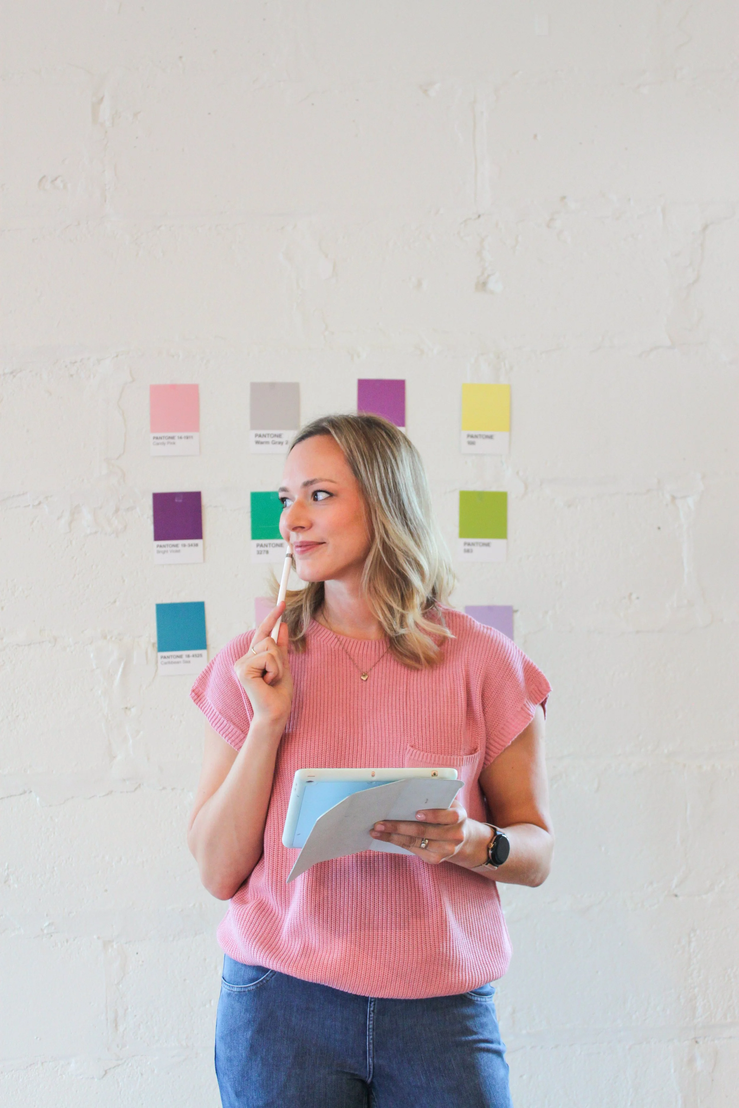 A woman with blonde hair wearing a pink top and blue jeans, holding a tablet and a pen, standing in front of a wall with colorful Pantone color swatches.