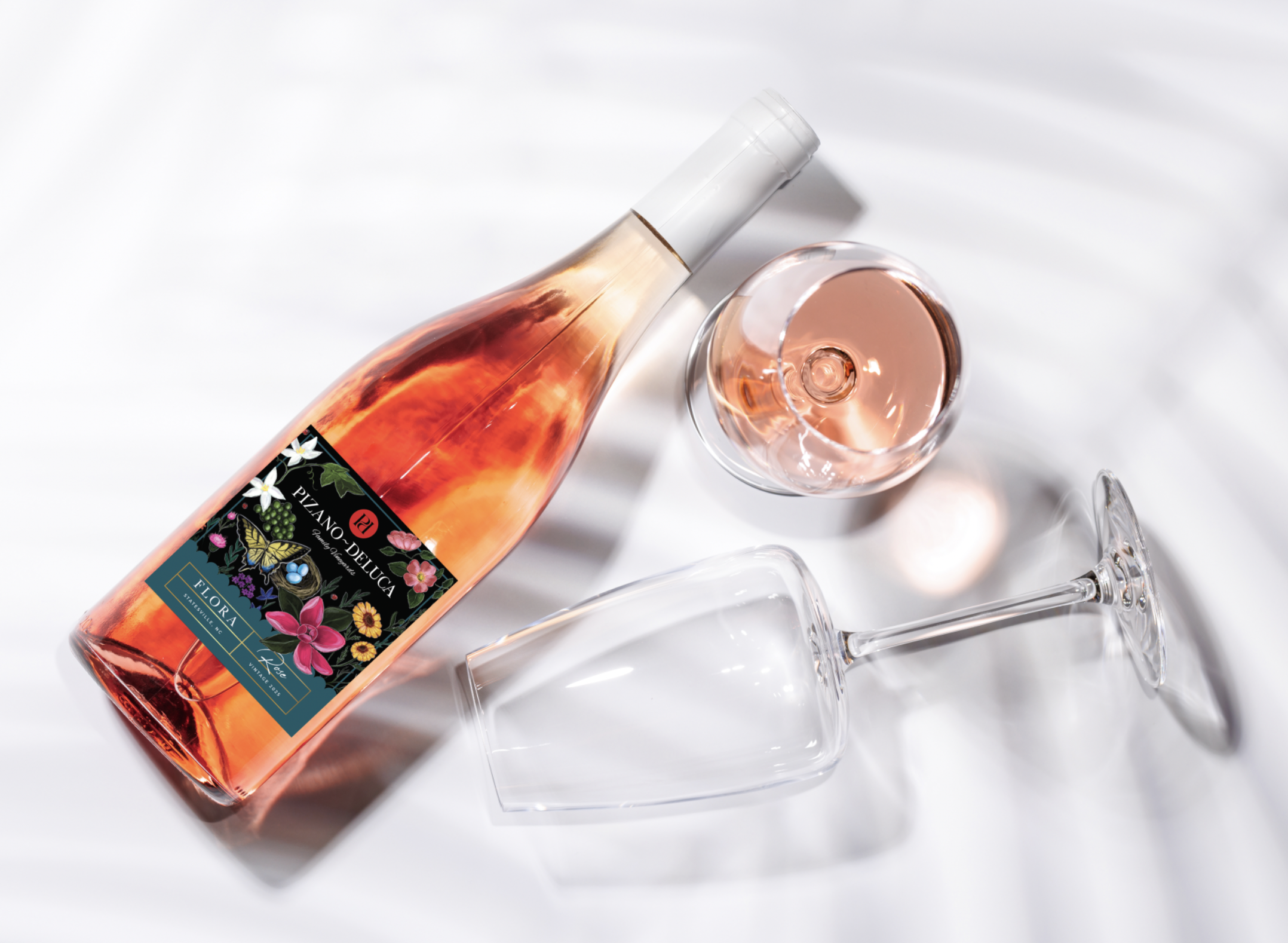 A bottle of rosé wine with a floral label, a filled wine glass, and an empty wine glass on a white surface.