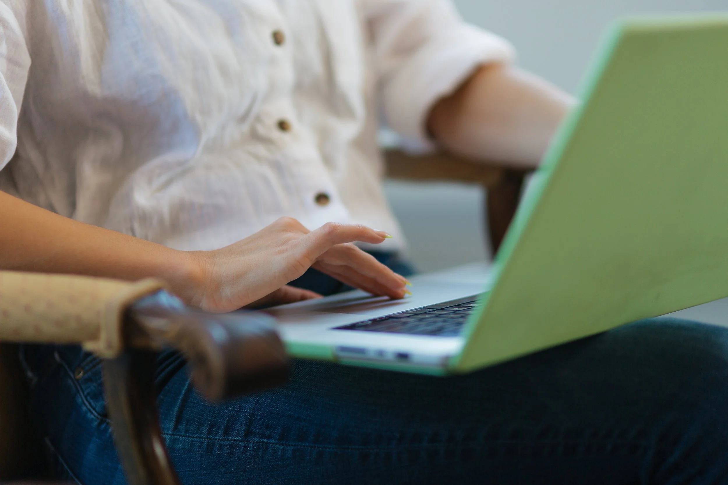 Person using a green laptop, sitting on a chair, wearing a white shirt and blue jeans.