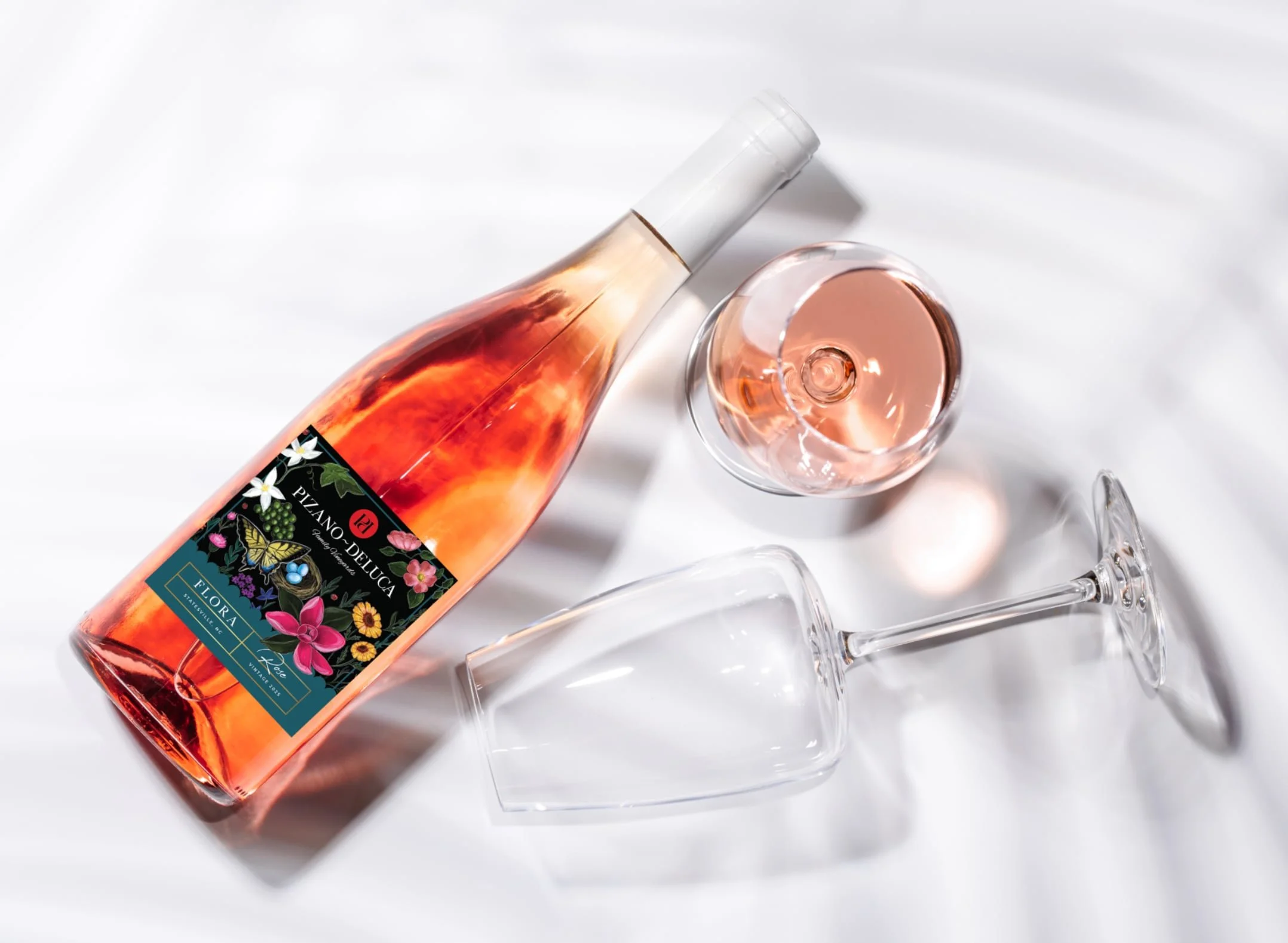 A bottle of rosé wine with a floral label, a filled wine glass, and an empty glass on a white surface with soft shadows.