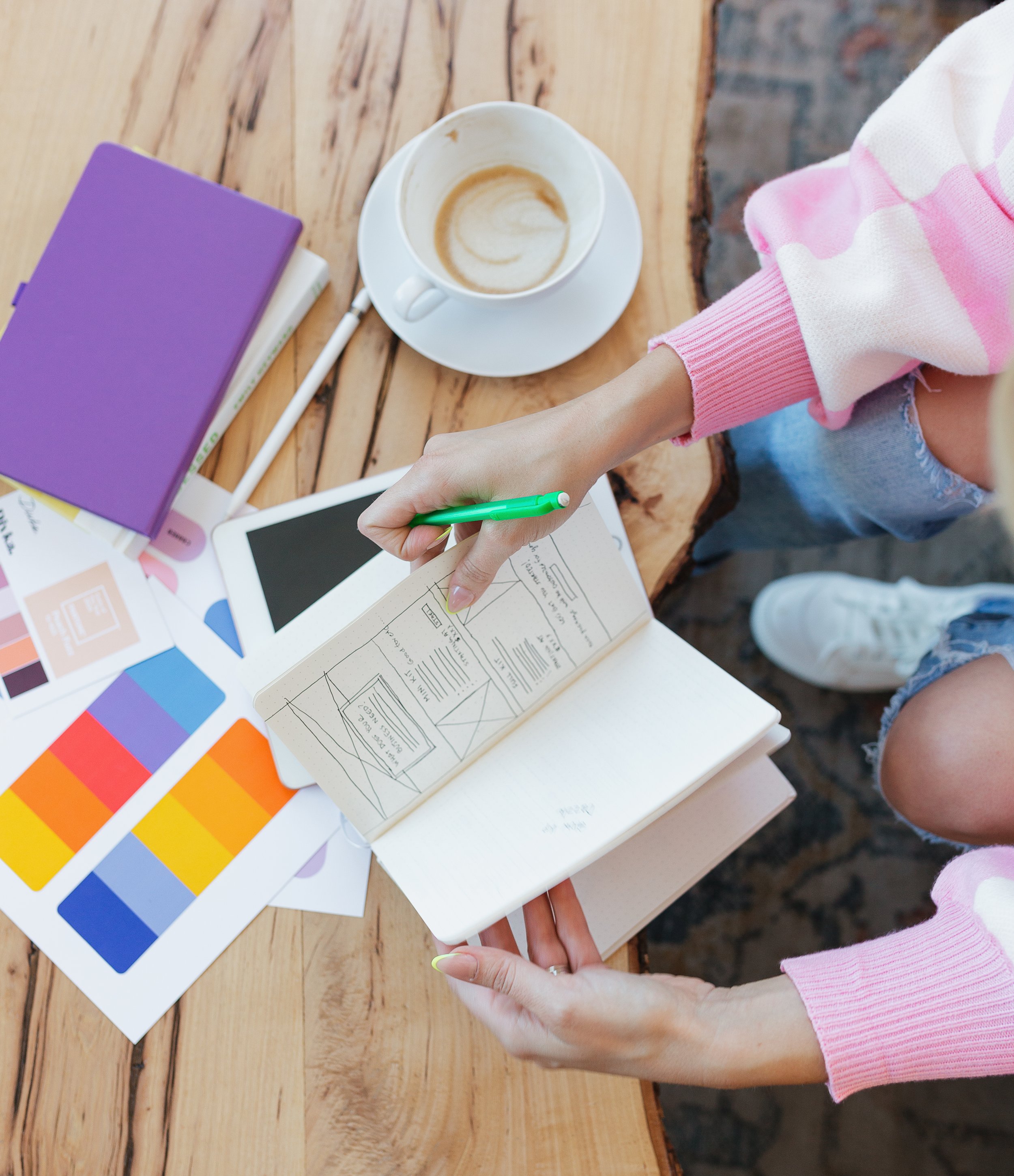A person is sitting at a wooden table with color swatches, a purple notebook, and a white tablet. They are using a green pen to sketch or write in a notebook. There is a coffee cup with a saucer and some latte art on the table.