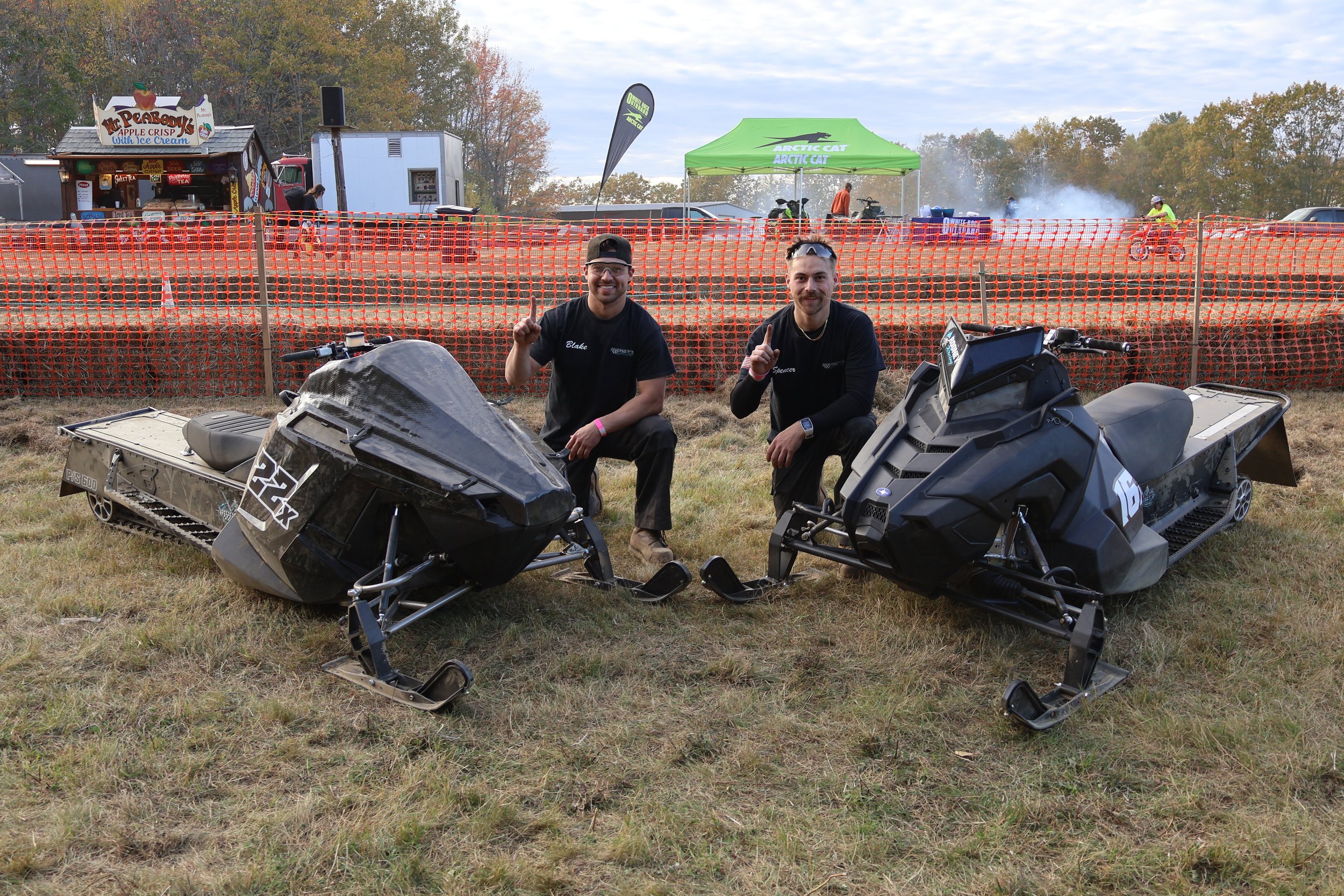 Two men kneeling on grass with snowmobiles in front of orange safety fencing at an outdoor event. One man is on the left and the other on the right, both smiling and giving a thumbs-up. In the background, there are tents, trailers, and several people.