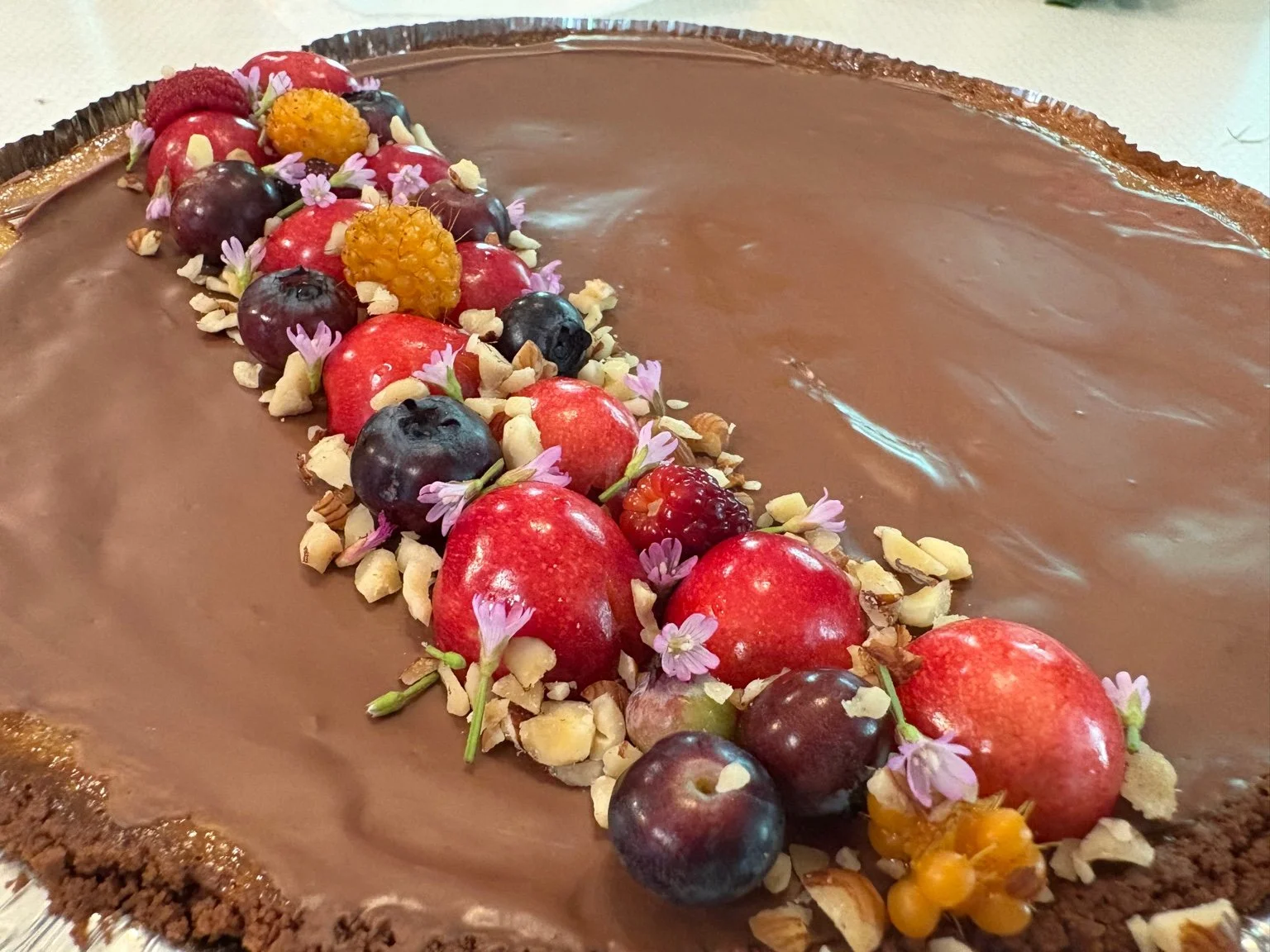 Chocolate tart with a variety of fresh berries, nuts, and small flowers on top.