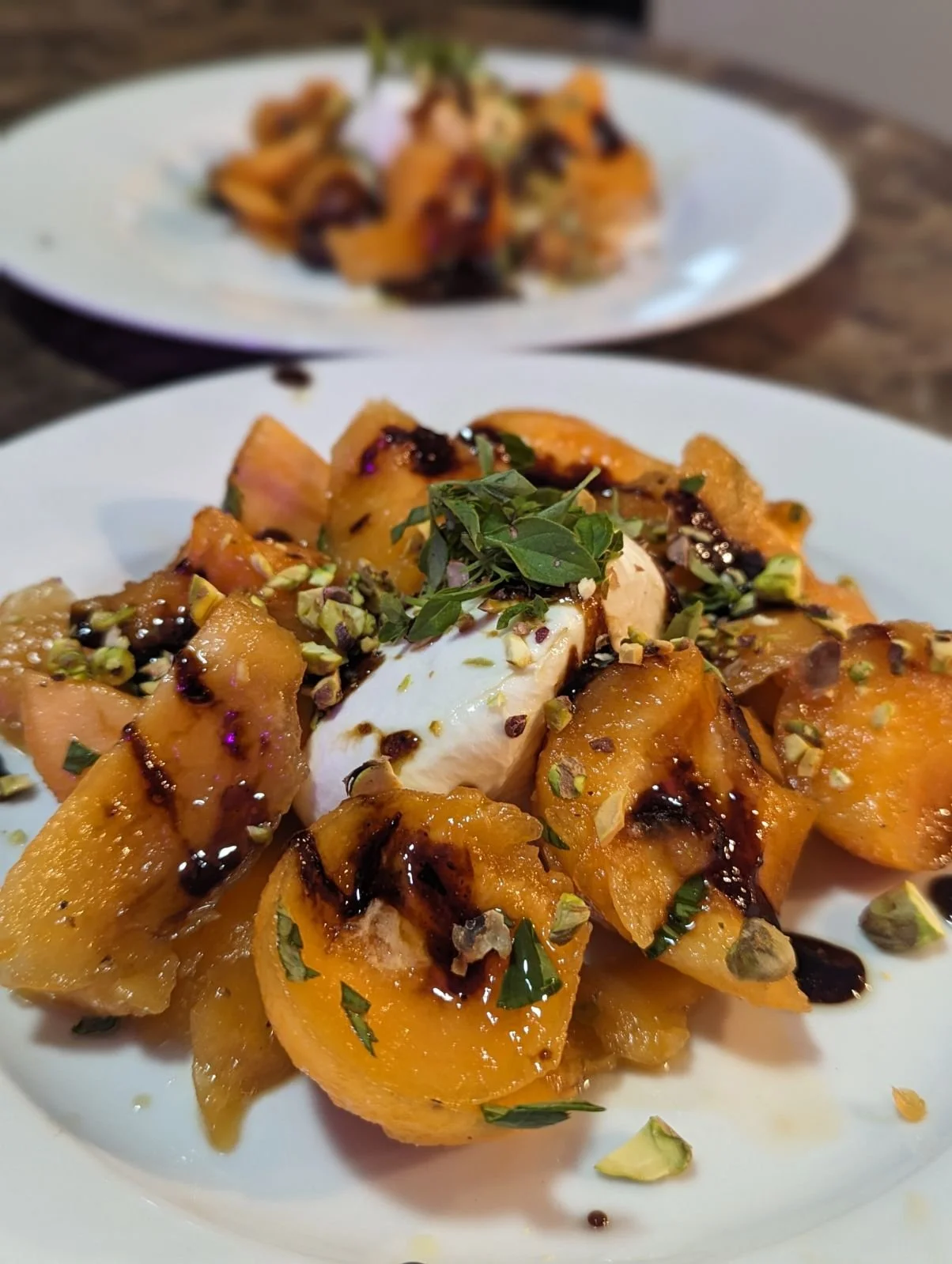 Close-up of a dish with sliced peaches, pistachios, herbs, and a dollop of cream, drizzled with syrup, served on a white plate.