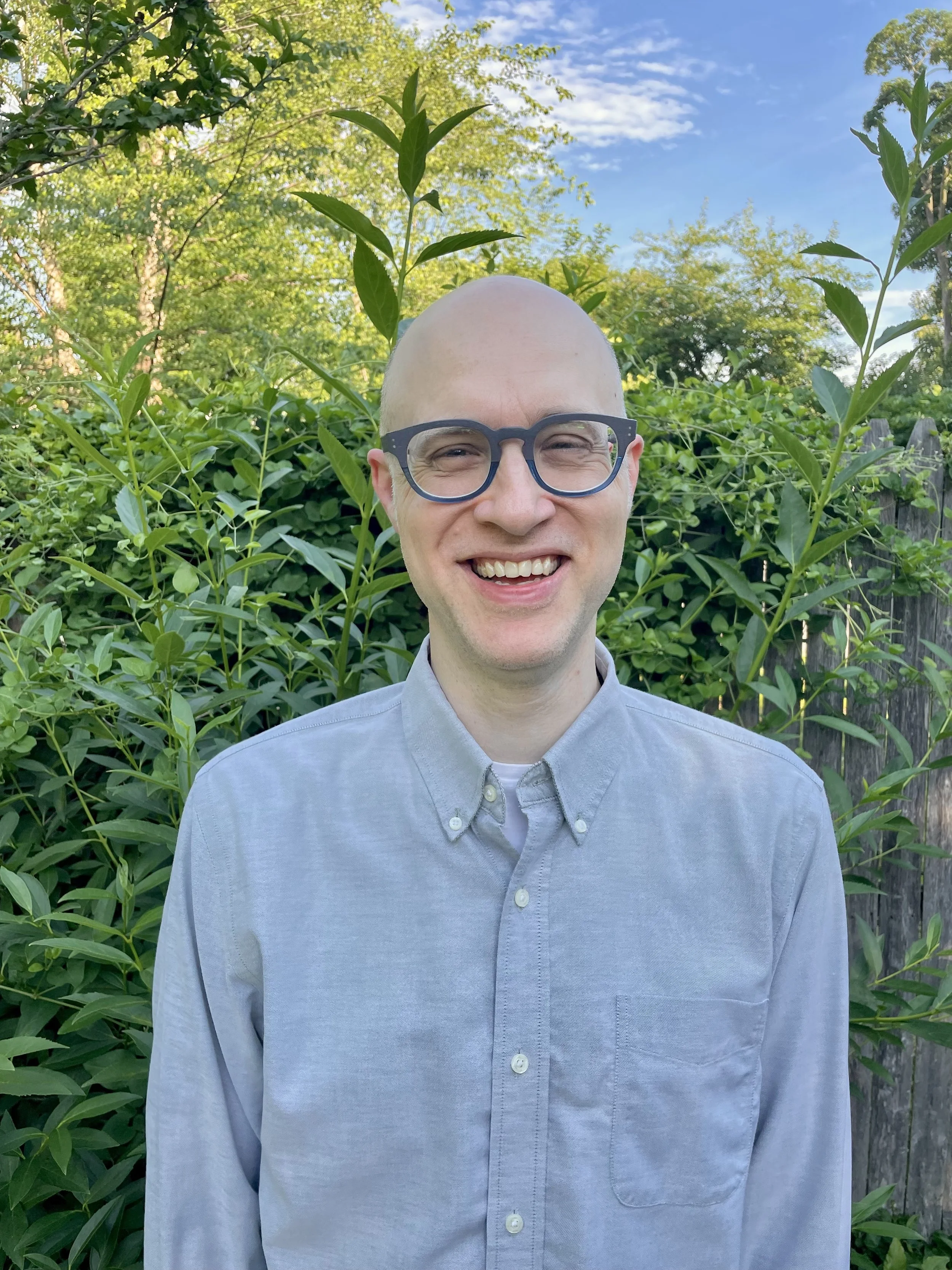 A smiling man with a bald head, wearing glasses and a light gray button-up shirt, standing outdoors in front of green foliage and trees on a sunny day.