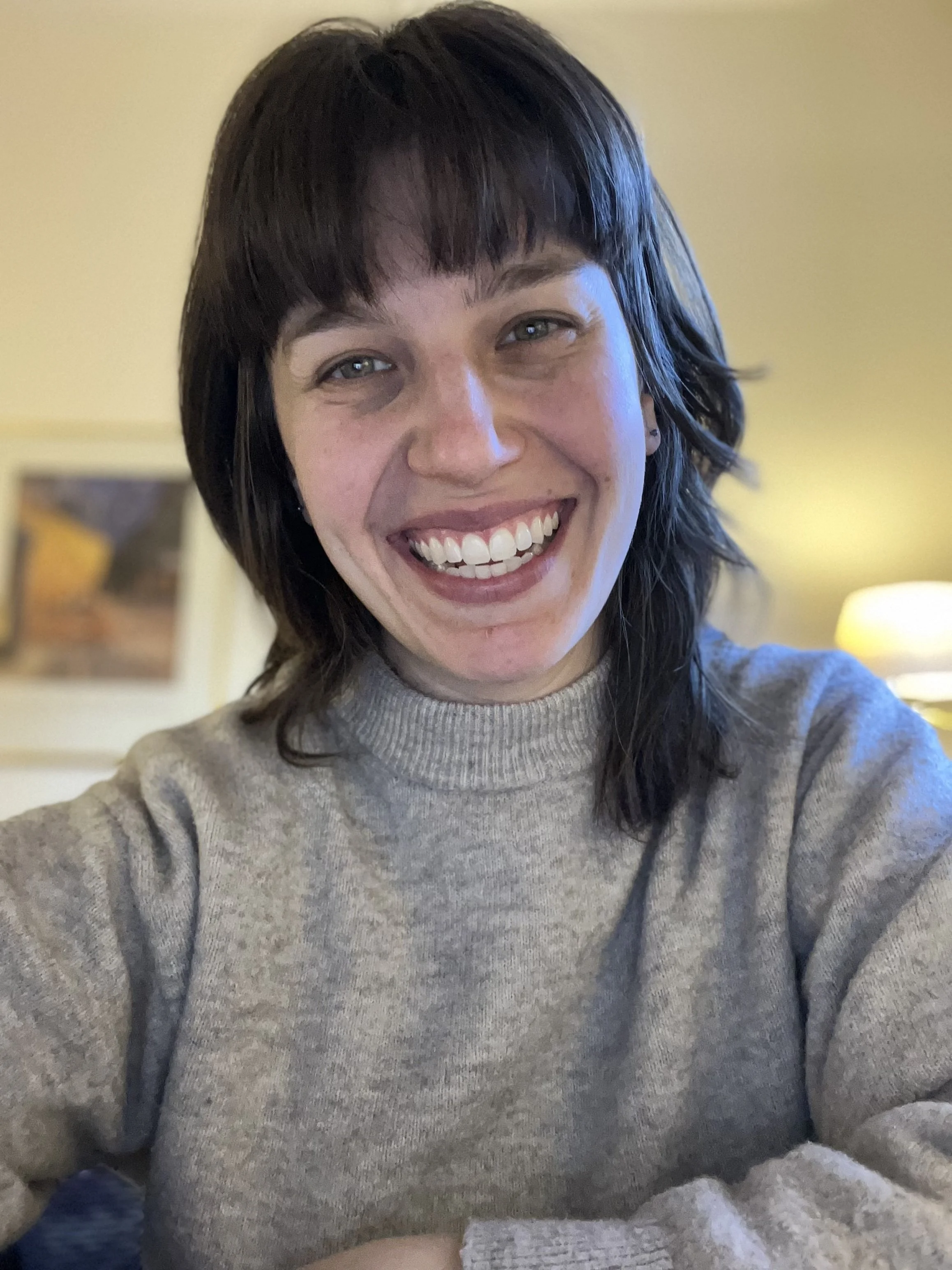 A woman with short dark hair, smiling, wearing a gray sweater, sitting in a warmly lit room with a blurred painting and lamp in the background.